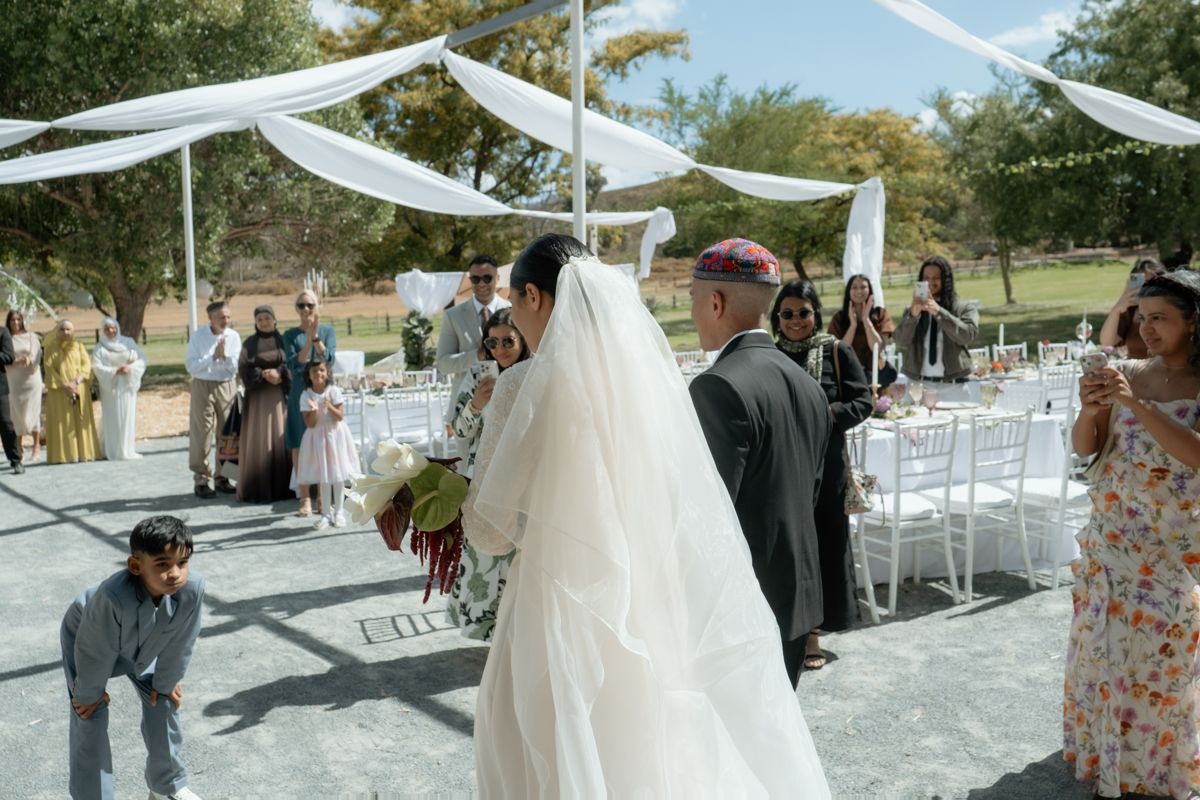 Bride in white veil walks down aisle at outdoor wedding ceremony with white draped canopy and guests.