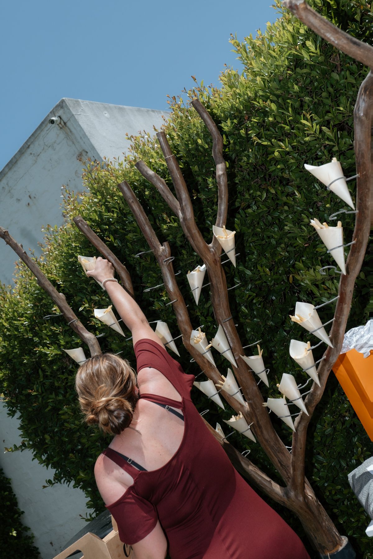 Woman in red dress arranging paper cone favors on a decorative bare branch display against a green hedge backdrop.
