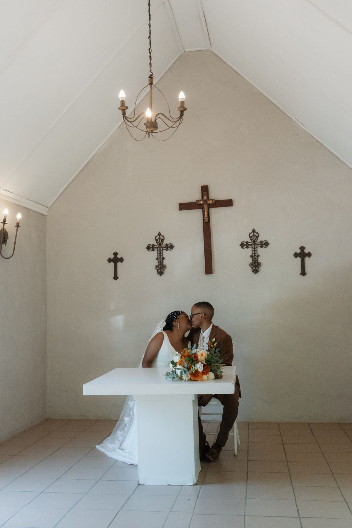Newlywed couple shares a kiss at a white altar inside a small chapel decorated with wooden crosses.