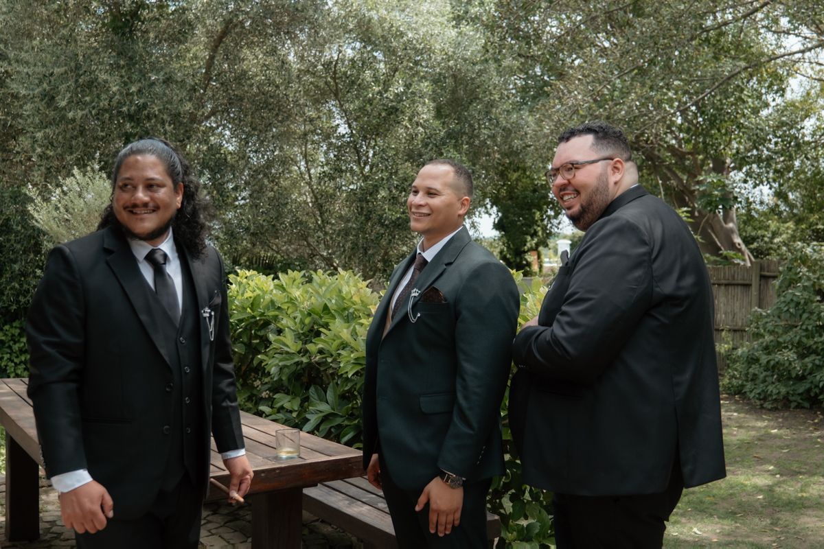 Three men in dark suits smile and chat outdoors in a garden setting before a wedding ceremony.