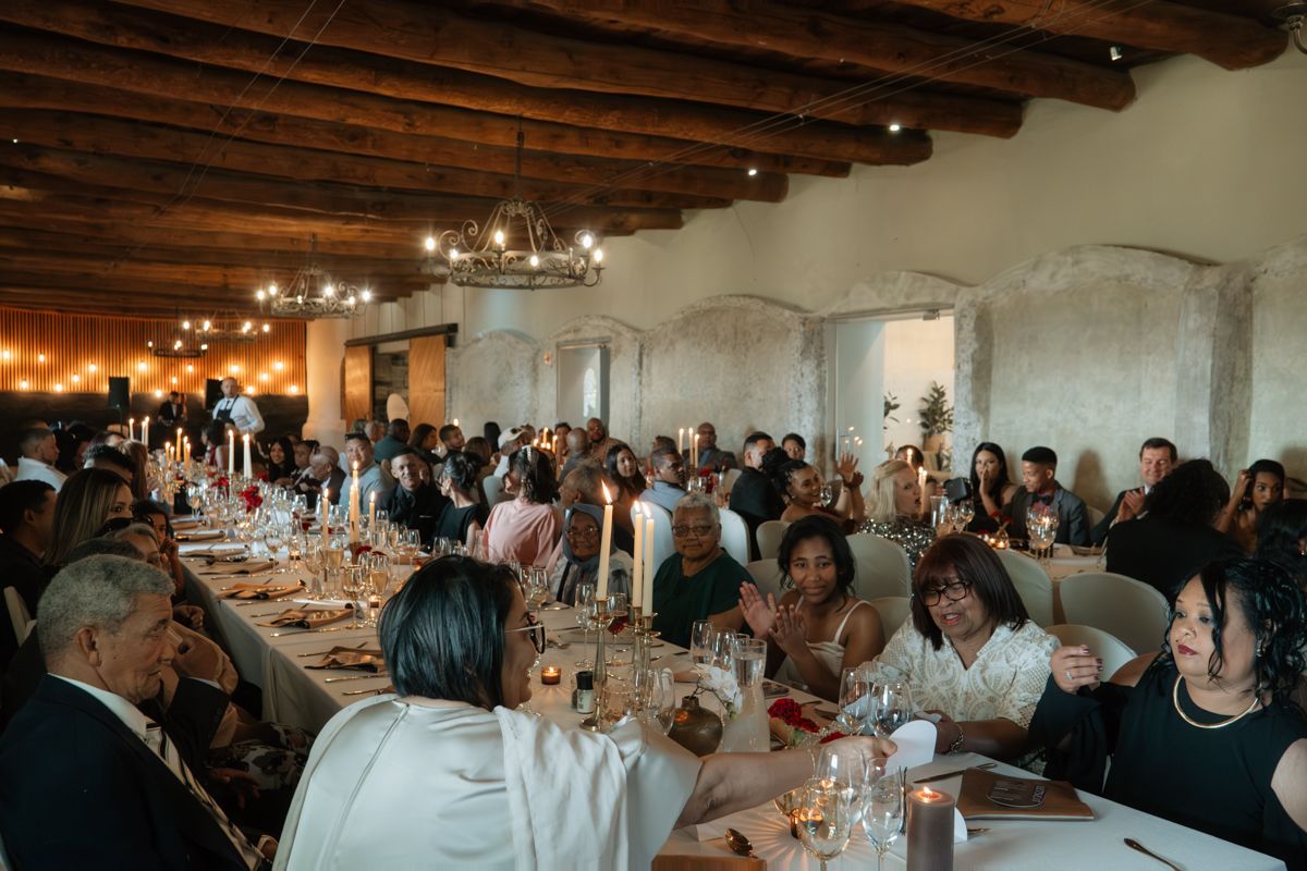 Large wedding reception dinner with guests seated at long tables in a rustic venue with wooden beam ceilings and candlelight.