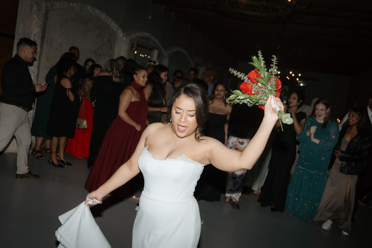 Joyful bride in strapless white gown raises her bouquet while dancing at her wedding reception.