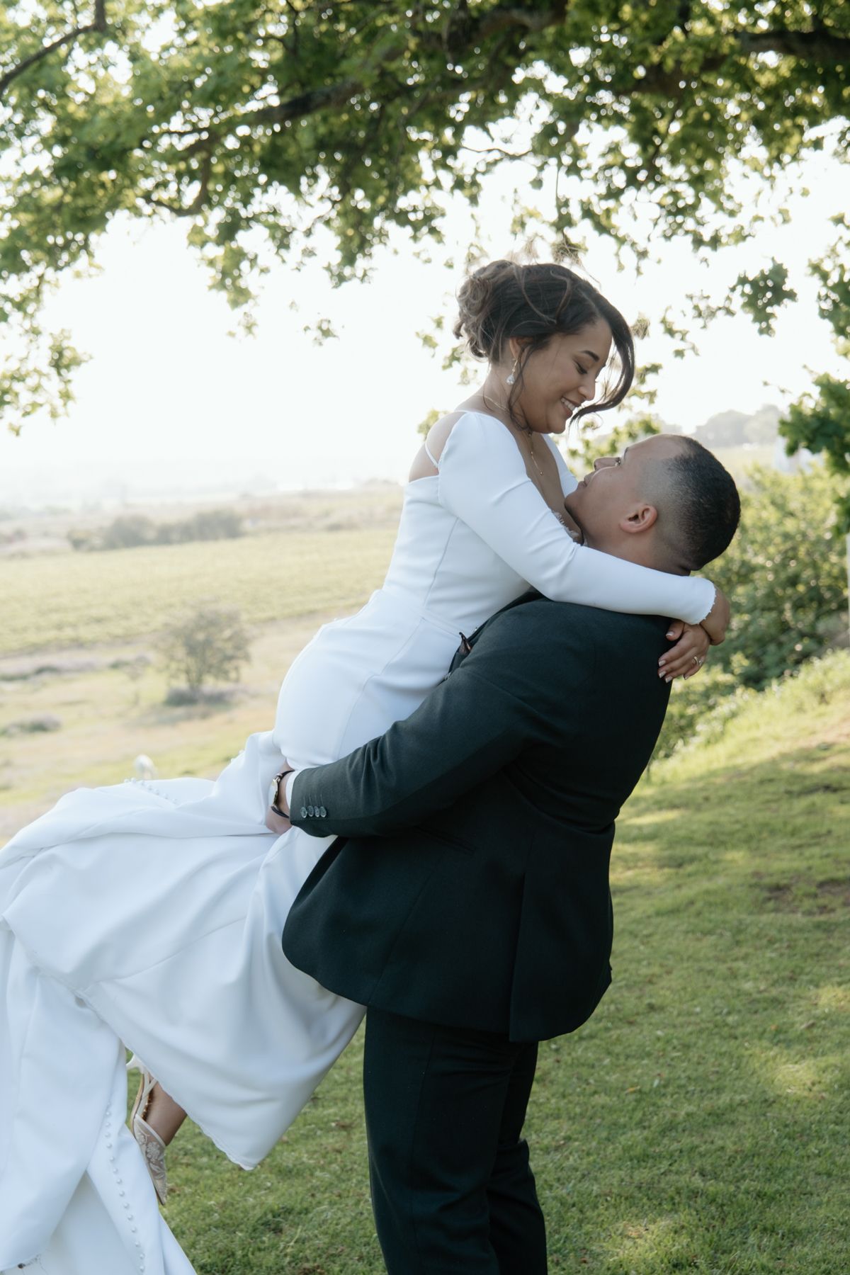Groom lifting smiling bride in white wedding gown beneath a tree in a scenic countryside setting.