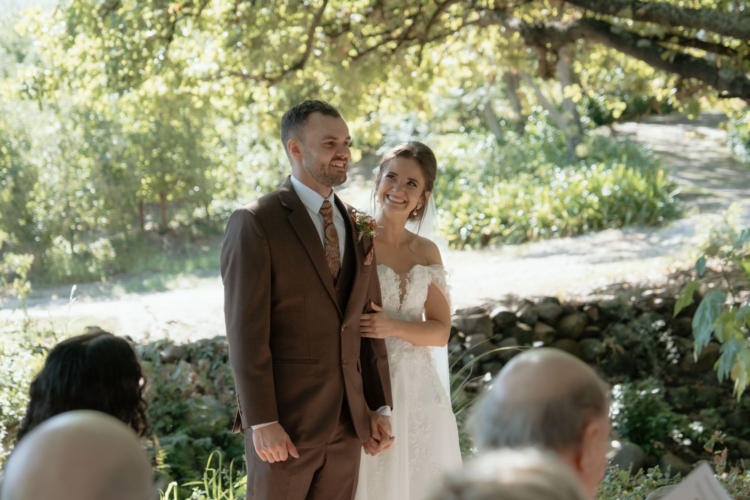 Bride and groom smile together at outdoor garden wedding ceremony surrounded by lush green trees.