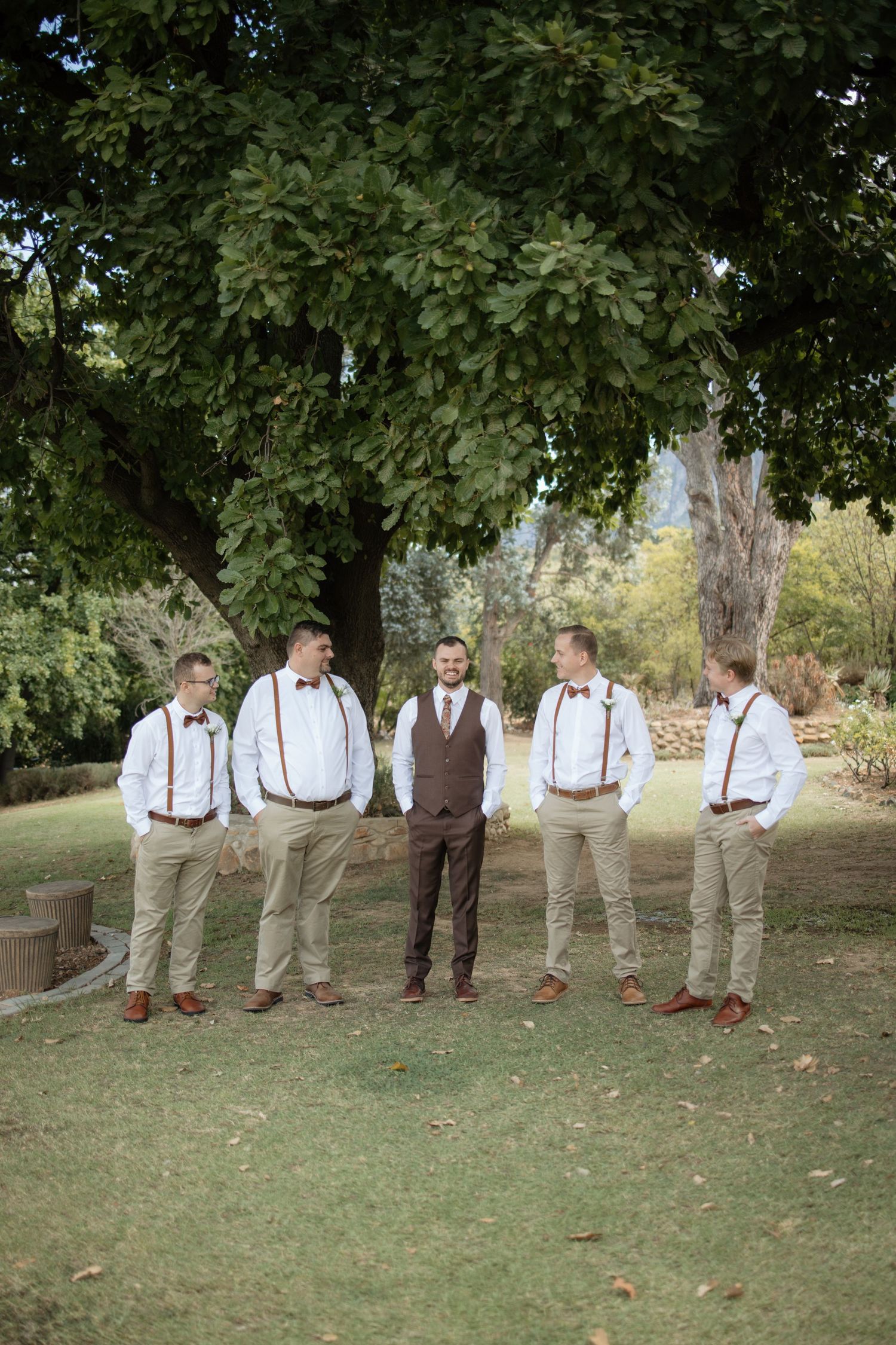 Groom and groomsmen in suspenders and bow ties pose outdoors under a large tree at a rustic wedding.