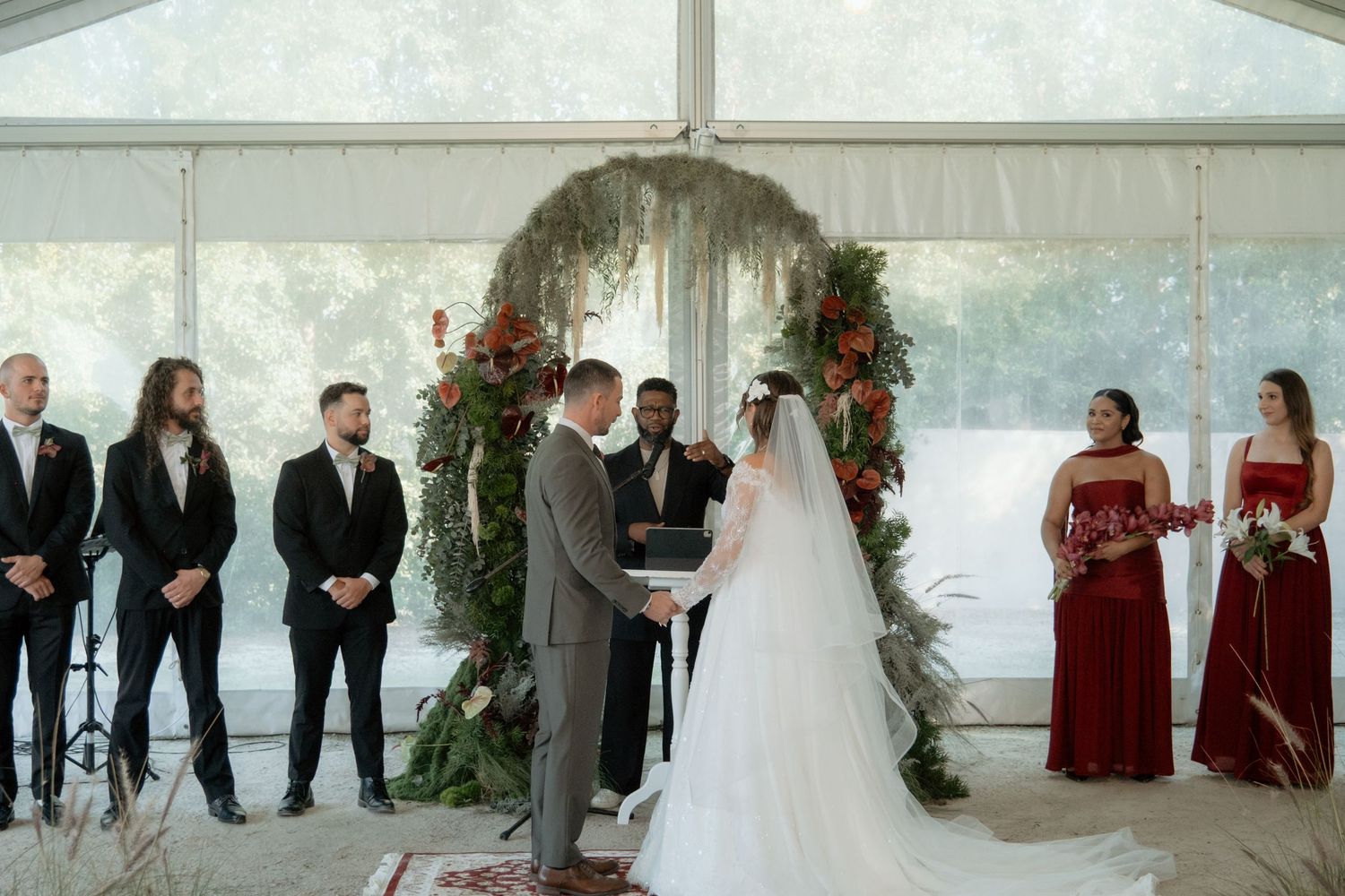 Couple exchanging vows at elegant tented wedding ceremony with floral arch, groomsmen, and bridesmaids in red dresses.
