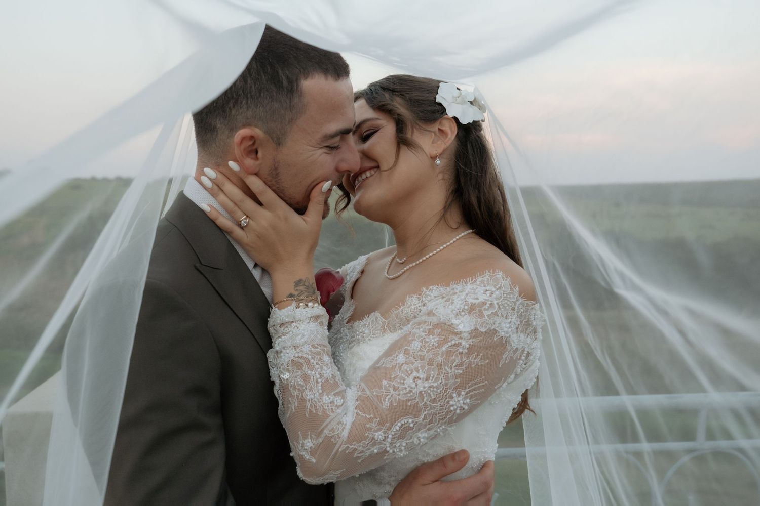 Romantic bride and groom almost kissing under flowing white veil at outdoor wedding photoshoot.