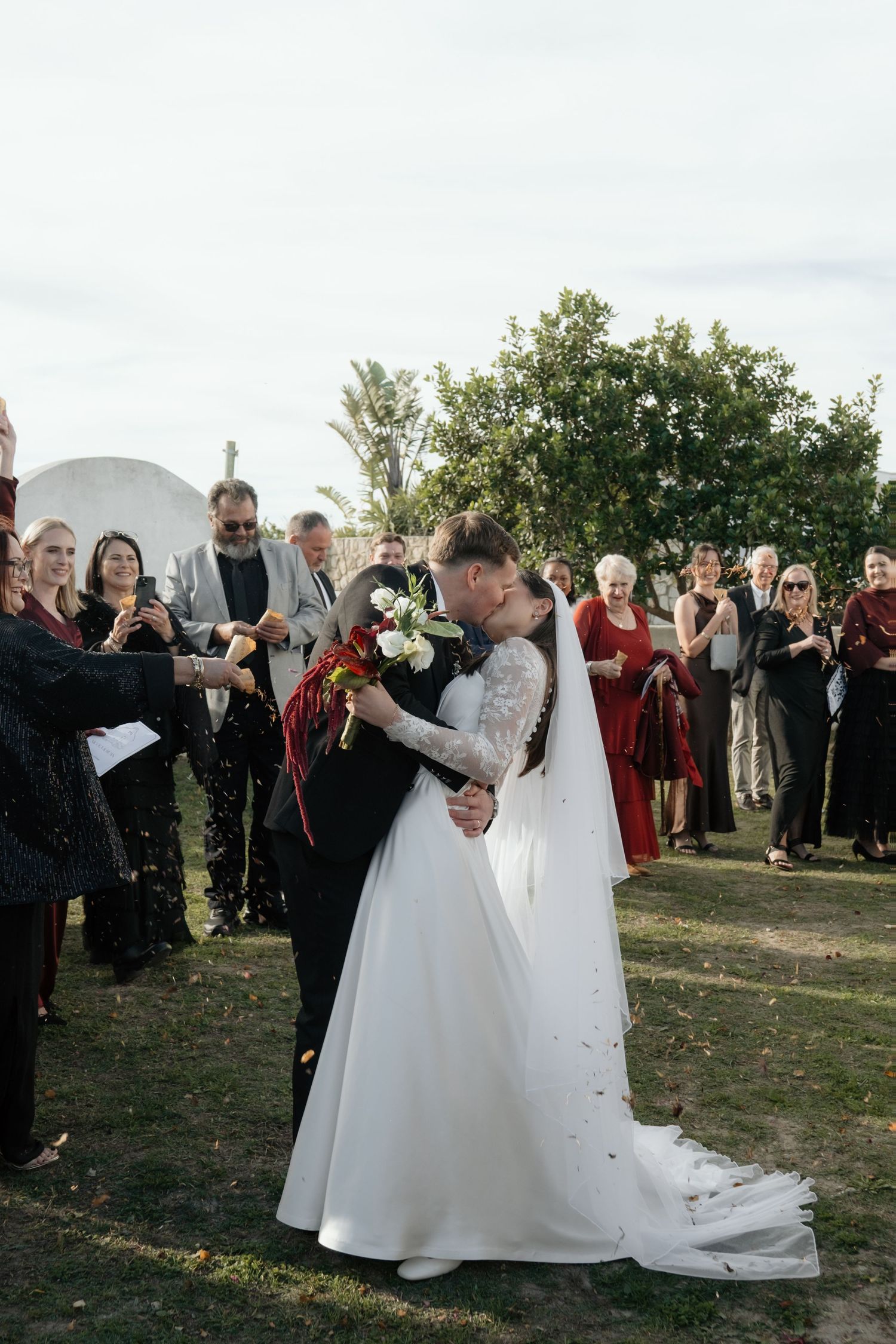 Bride and groom share first kiss as newlyweds while wedding guests celebrate with confetti outdoors.