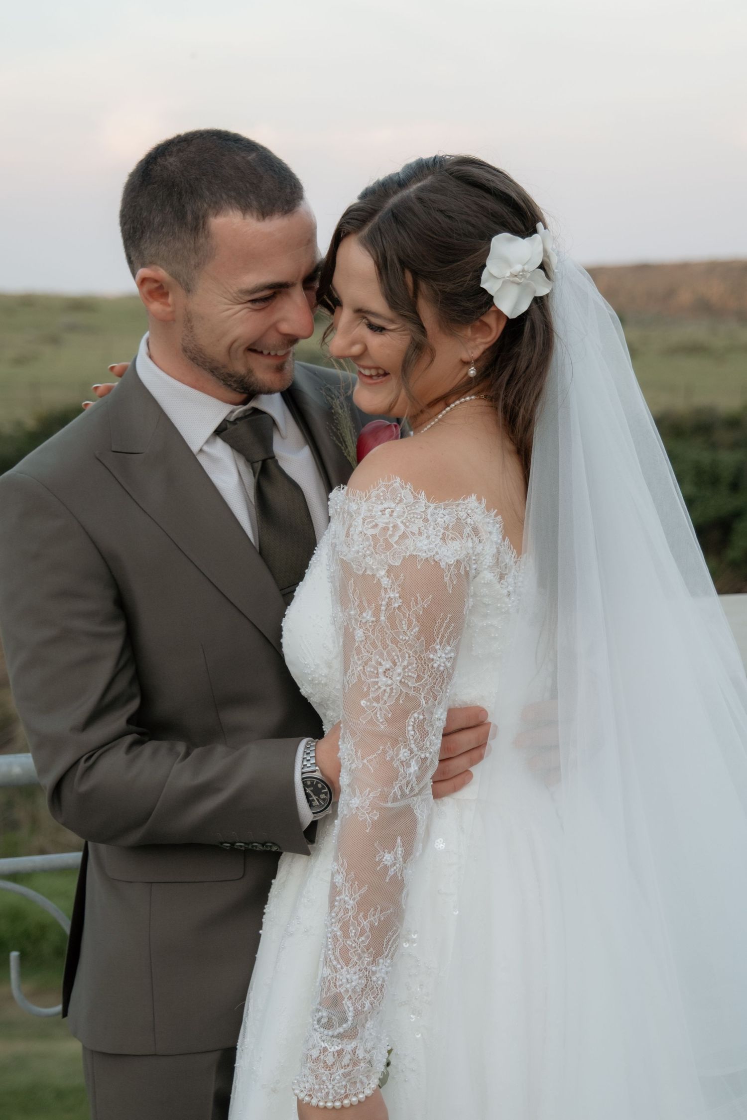 Smiling bride and groom embracing outdoors at dusk, bride in lace dress with veil.