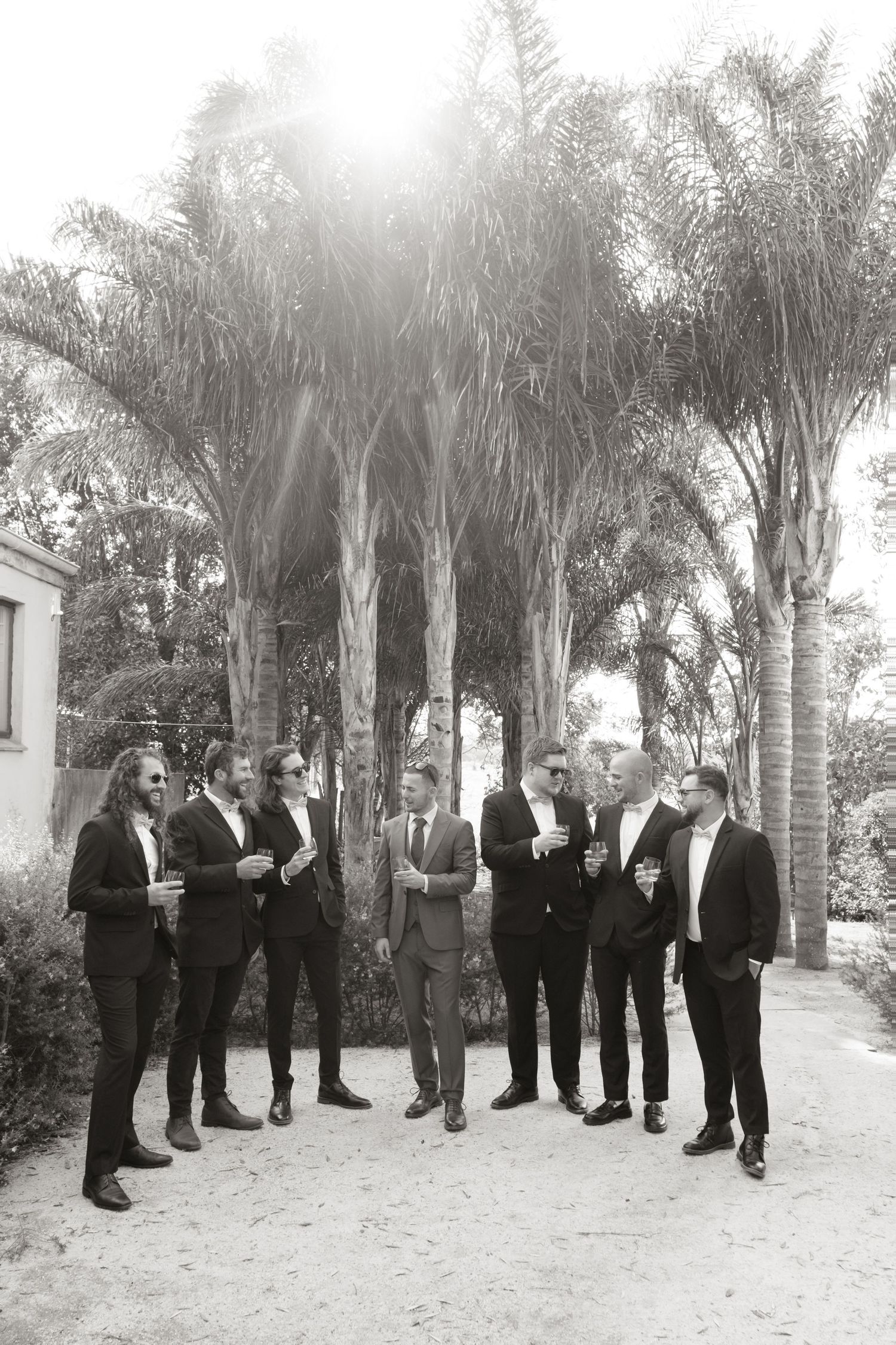 Black and white photo of groomsmen in suits gathered outdoors under palm trees at a wedding.