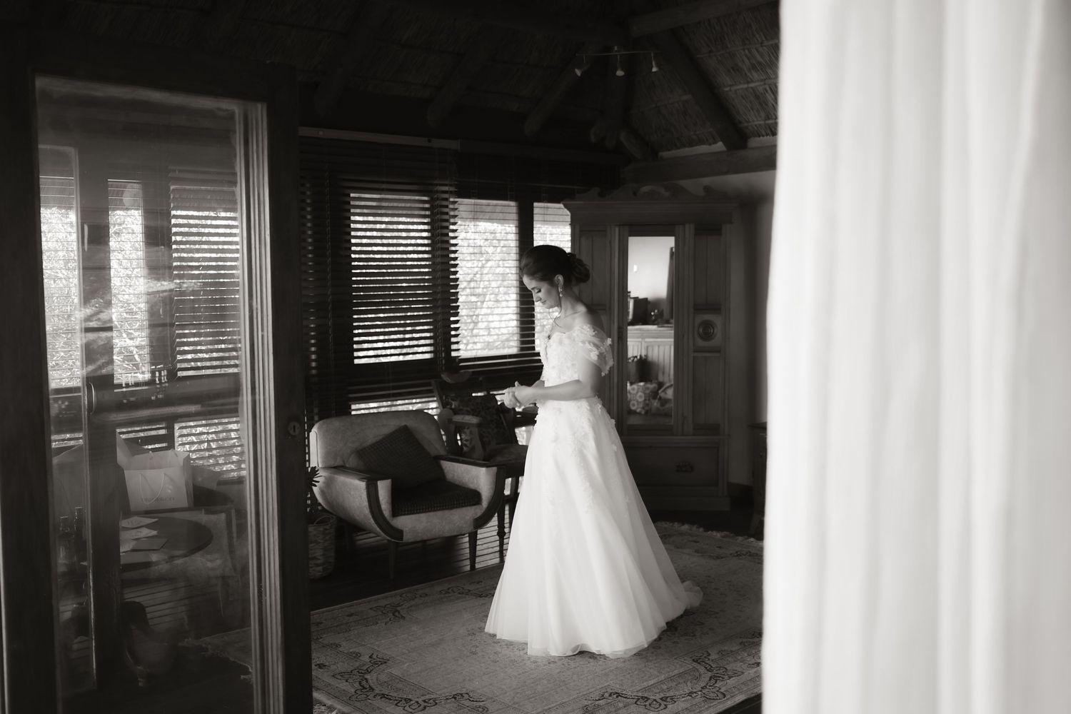 Black and white photo of a bride in a white gown standing by a window in an elegant room on her wedding day.