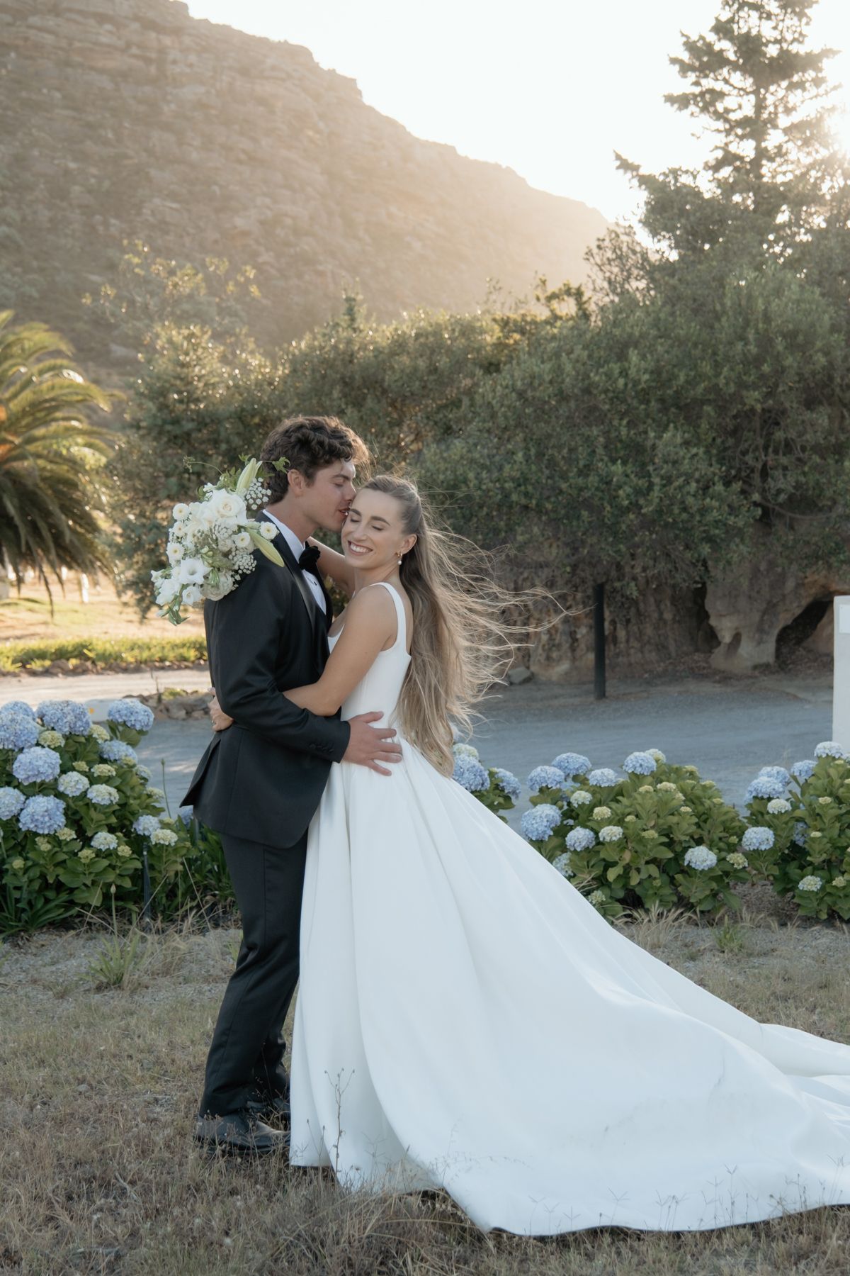 Bride and groom sharing romantic embrace in golden sunlight surrounded by hydrangeas and mountains.