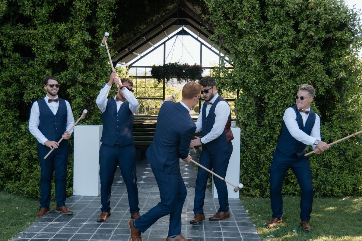 Groomsmen in navy suits playfully dueling with canes outside wedding venue.