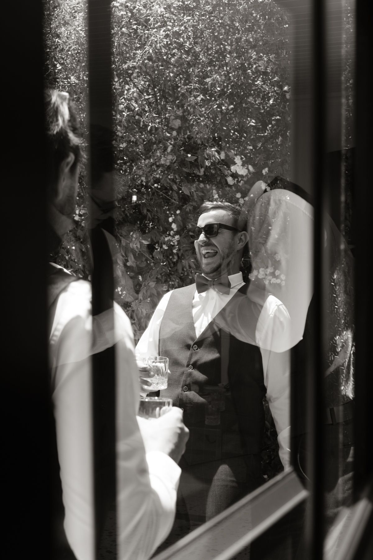 Black and white photo of a laughing groomsman in vest and bow tie enjoying wedding reception drinks.