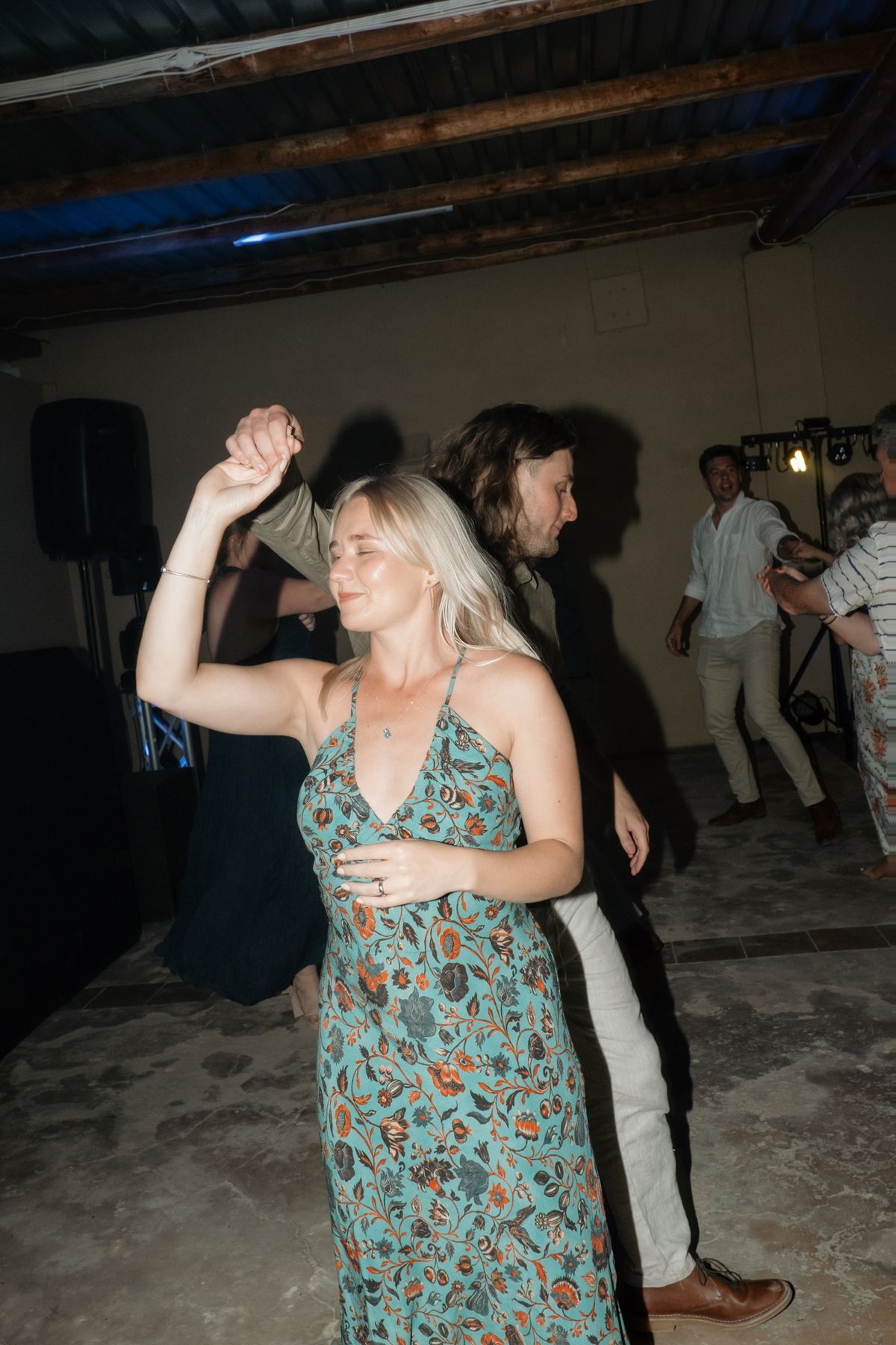Guest in floral maxi dress dancing joyfully at lively indoor wedding reception party.