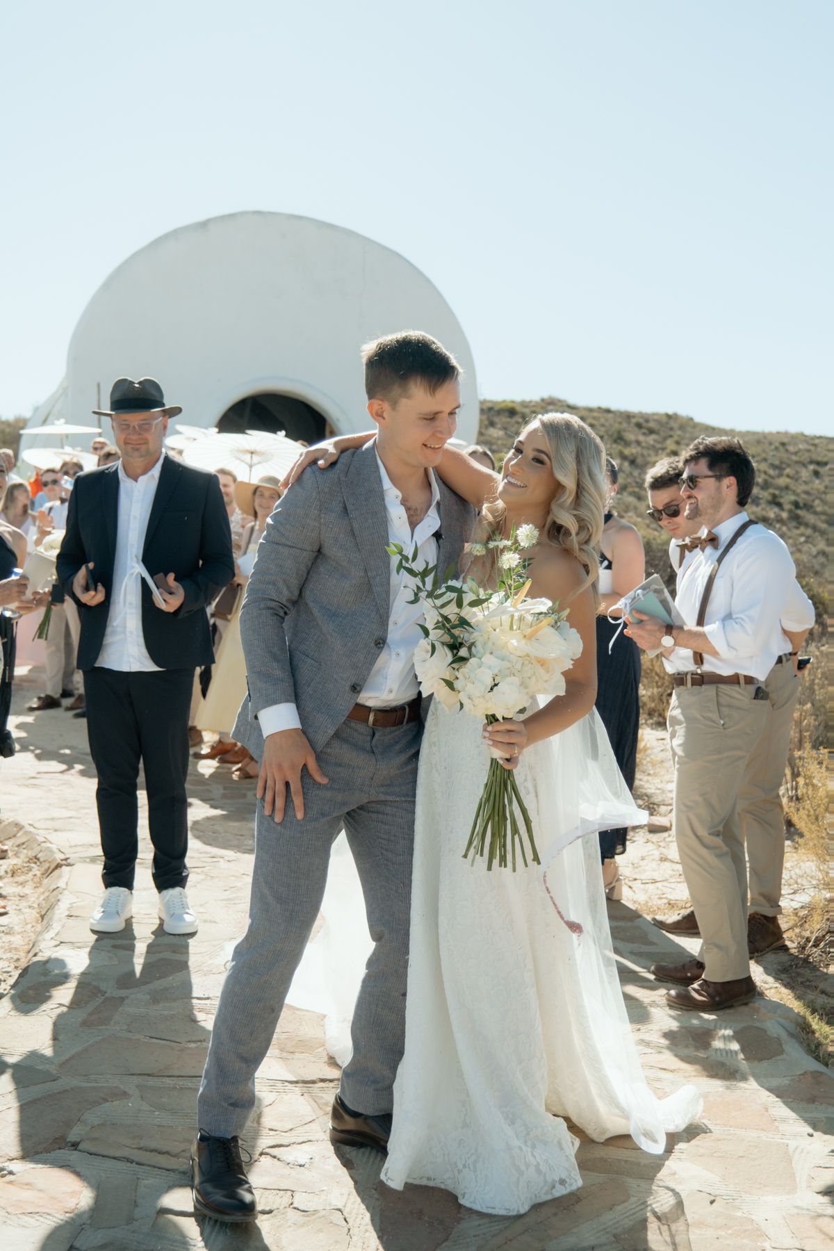 Bride and groom share a tender moment outside a white chapel while wedding guests celebrate around them.