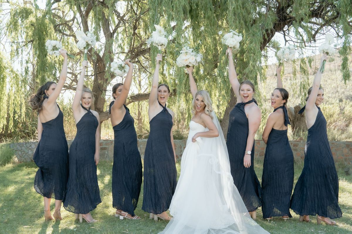 Bride and six bridesmaids in navy halter dresses raise bouquets joyfully under a blooming willow tree.