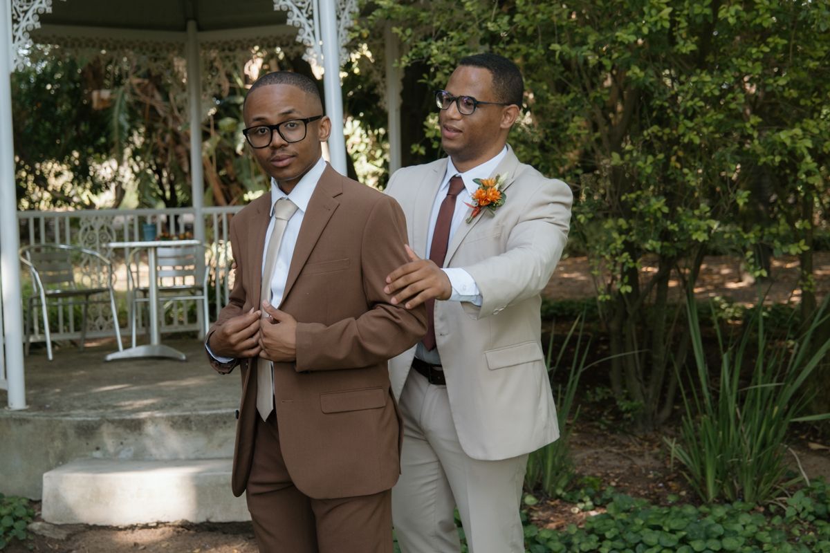 Two men in elegant suits pose outdoors near a gazebo, one adjusting the other's jacket at a formal event.