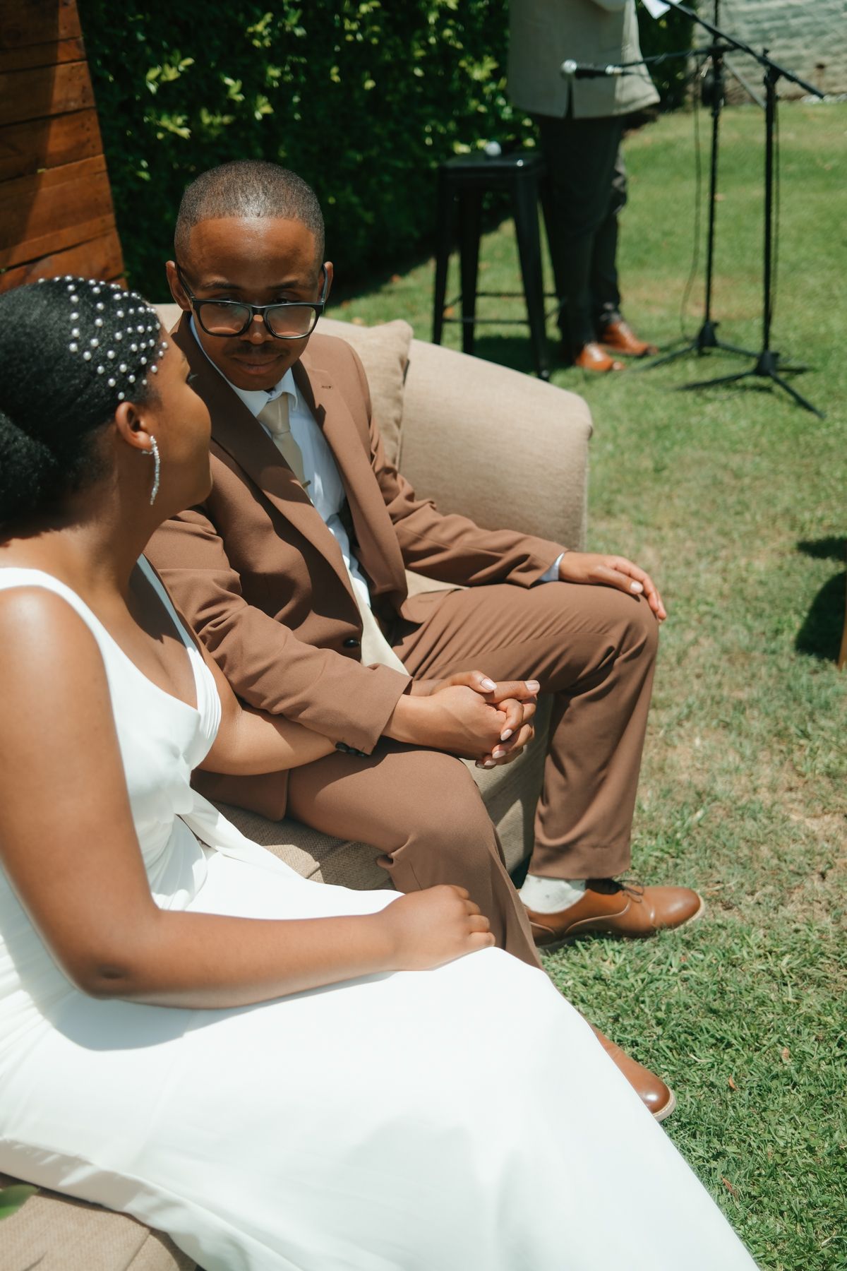 A Black couple in wedding attire holds hands outdoors, groom in brown suit with glasses, bride in white dress.
