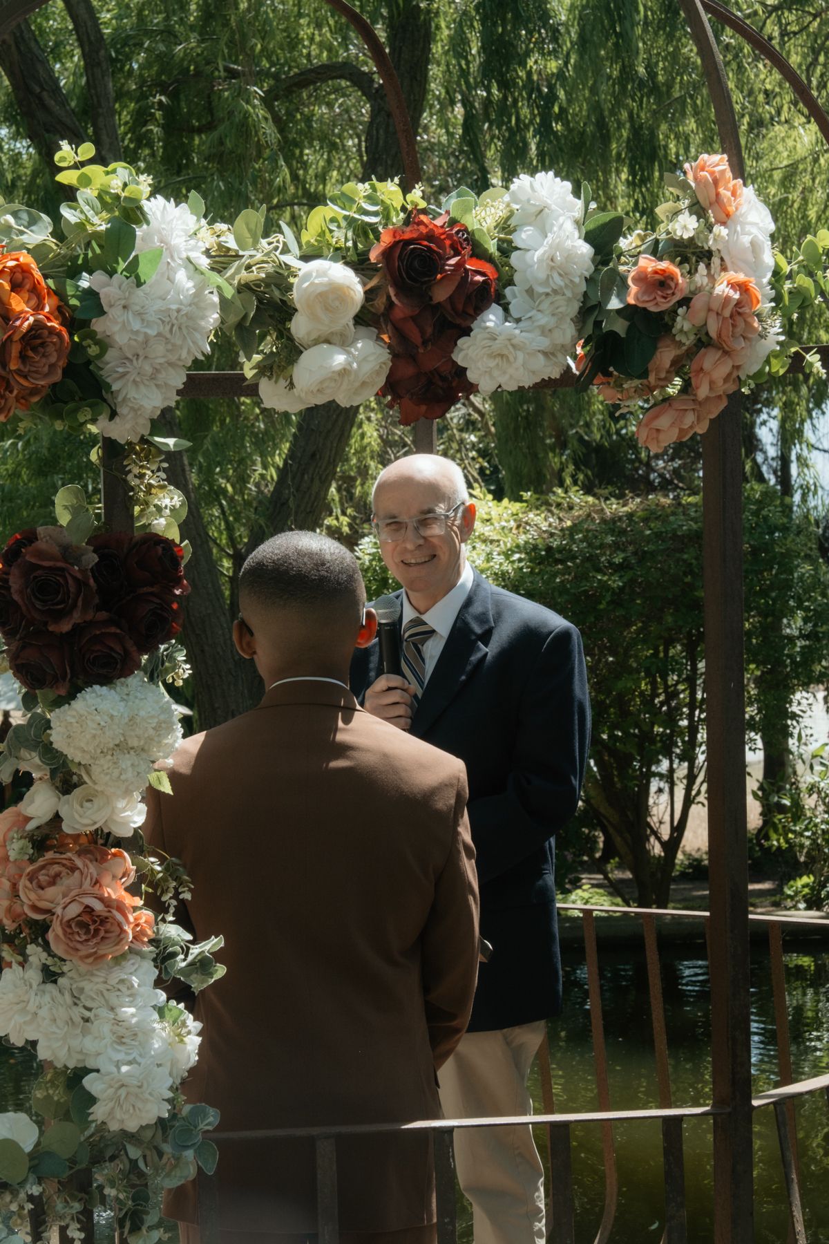 Groom in brown suit exchanges vows under floral arch with officiant at outdoor wedding ceremony.