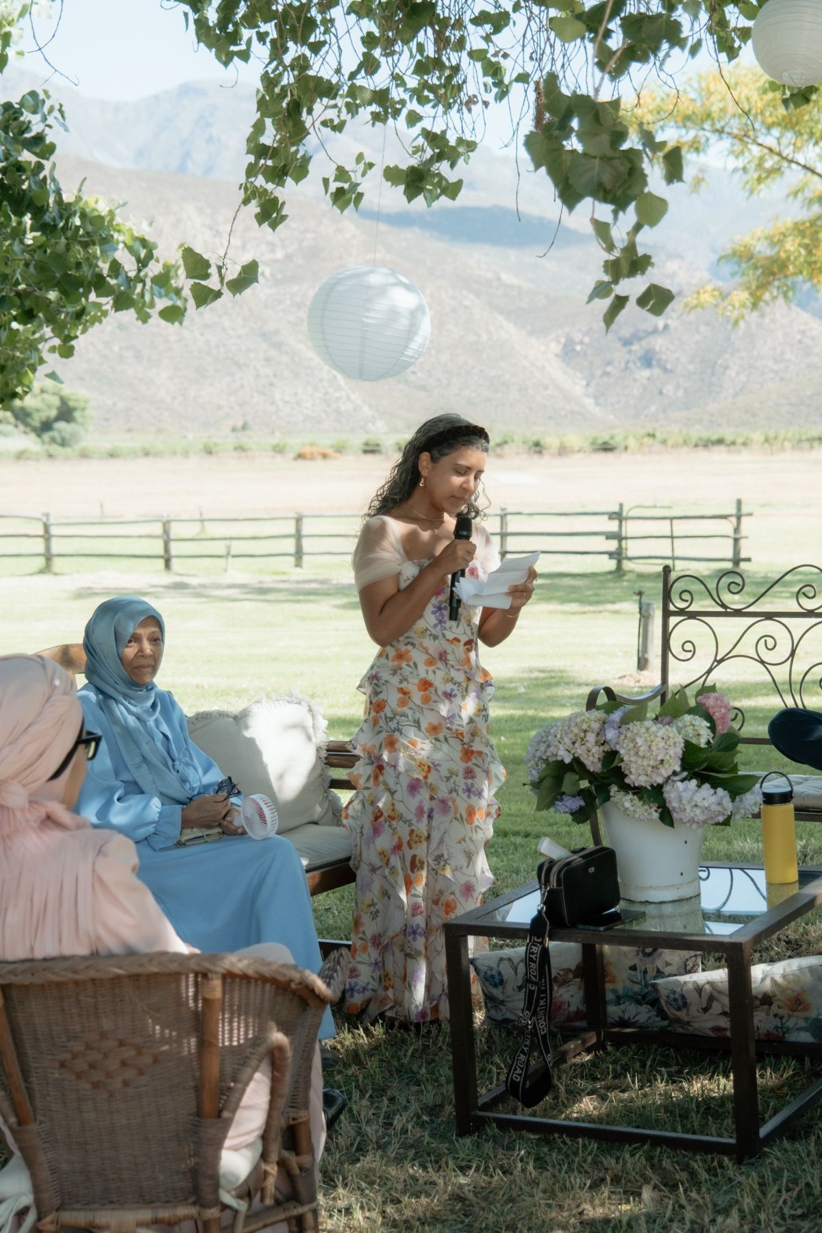 Woman in floral dress giving speech at outdoor wedding with mountain backdrop and balloon decorations.