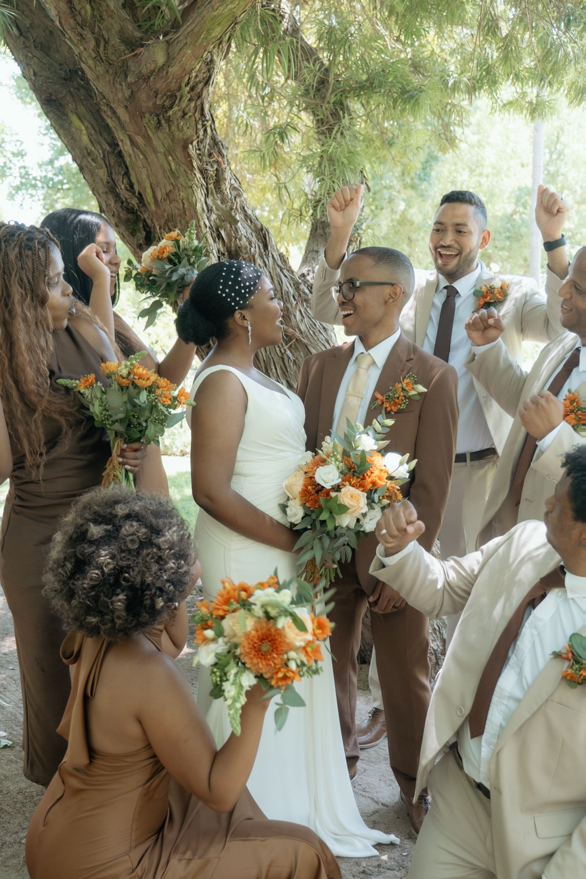 Joyful wedding party celebrating outdoors under a tree, bride in white holding orange floral bouquet with bridesmaids in brown dresses.