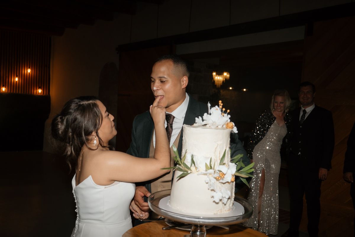 Bride feeds wedding cake to groom during cake cutting ceremony beside a white floral-decorated tiered wedding cake.