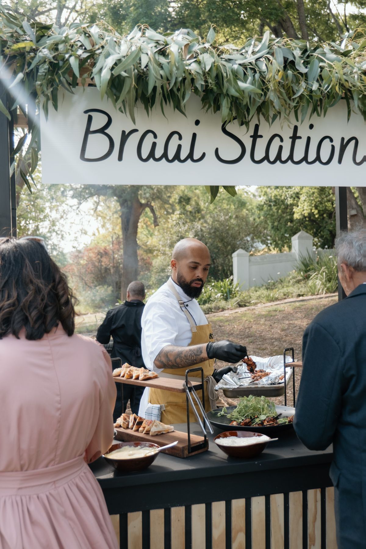 Chef serving guests at decorated Braai Station food station at outdoor wedding reception.