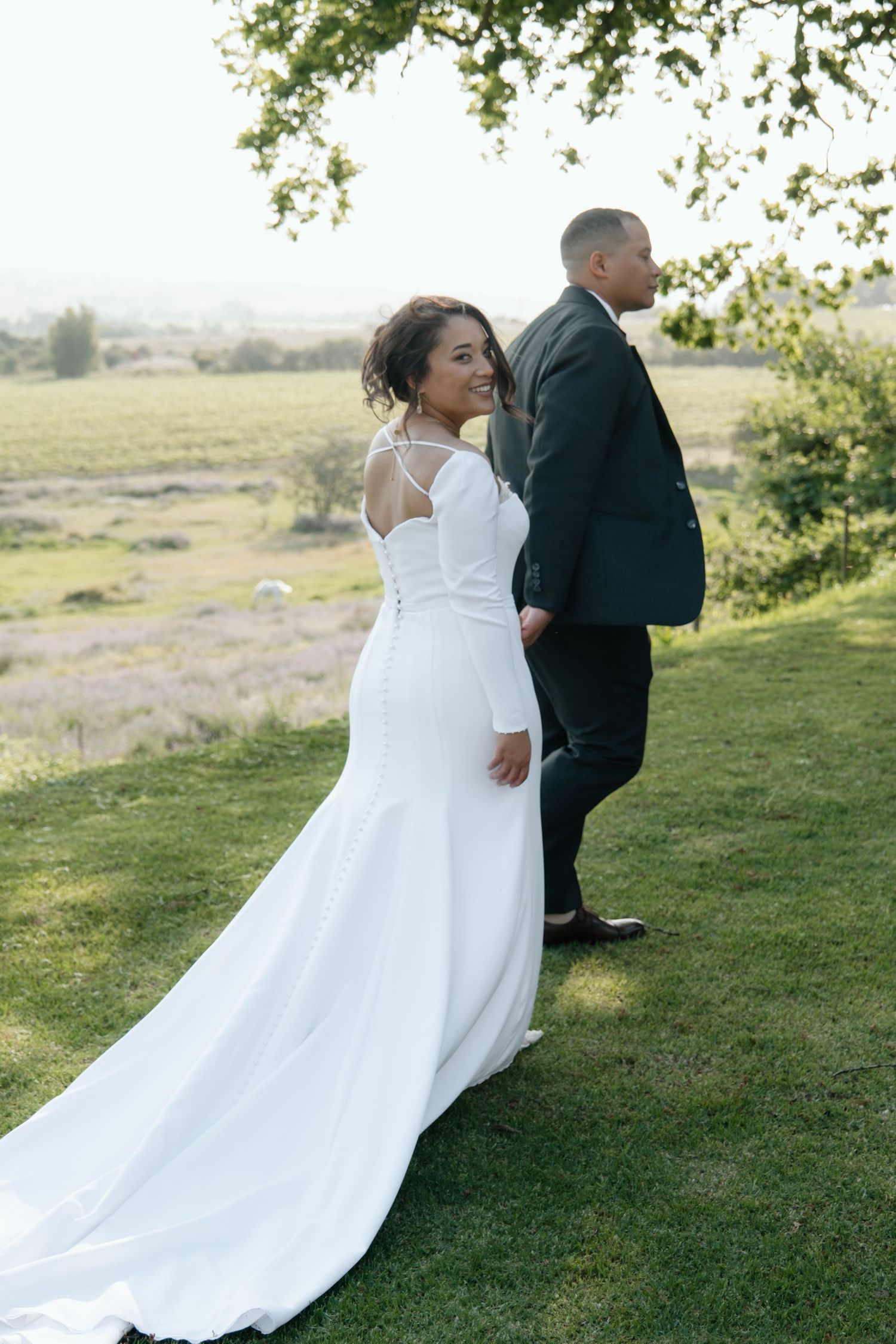 Bride in elegant white mermaid wedding dress with train smiles back at camera while walking with groom on green lawn.