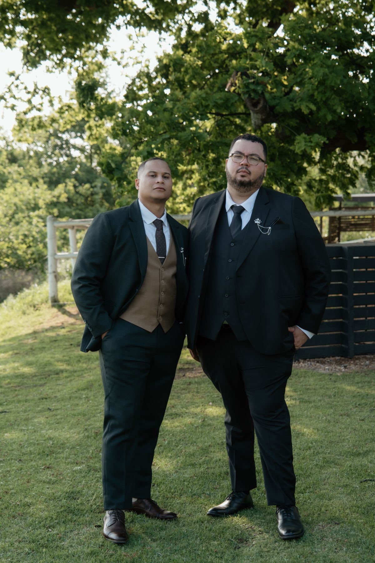 Two groomsmen in dark suits with waistcoats posing confidently outdoors under a leafy green tree.