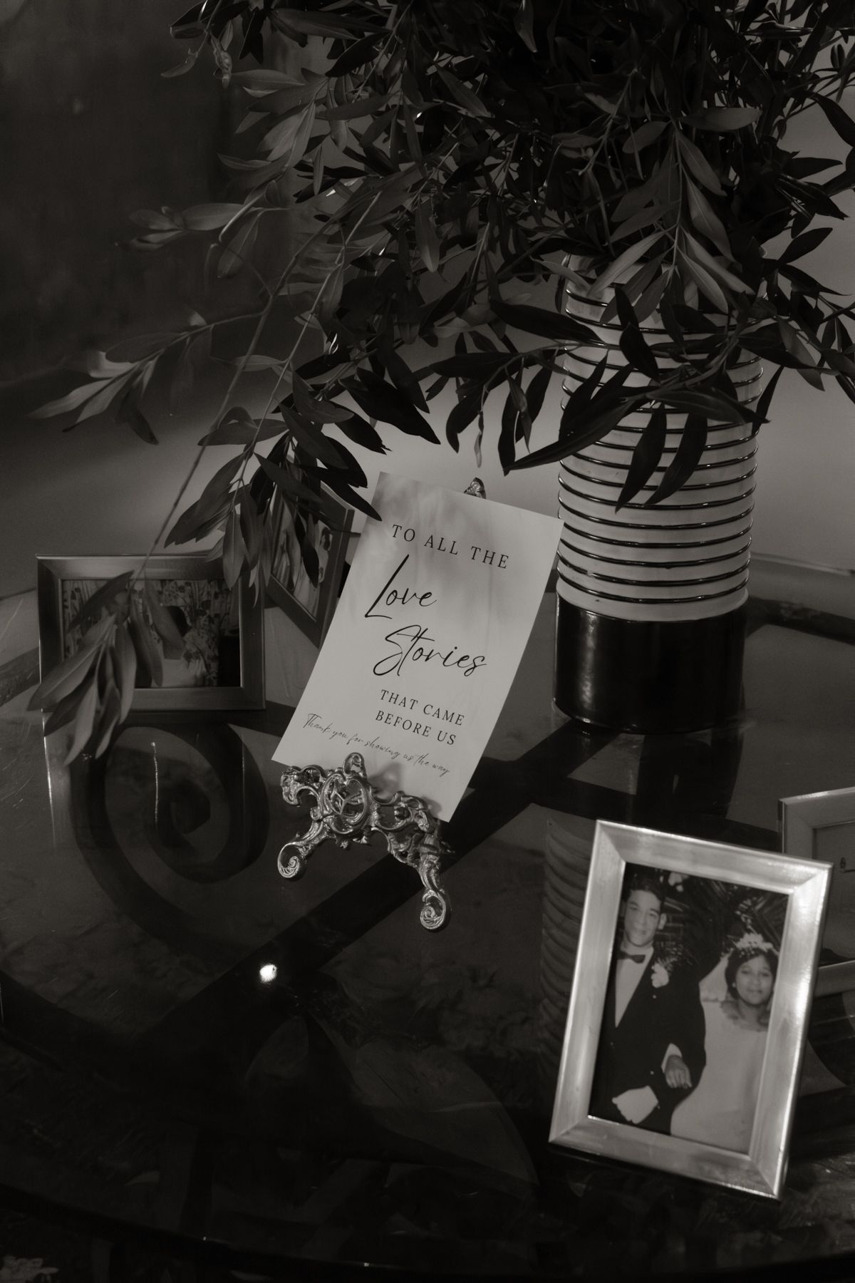 Black and white photo of a wedding memory table with framed photos, a love stories sign, and a striped vase with greenery.