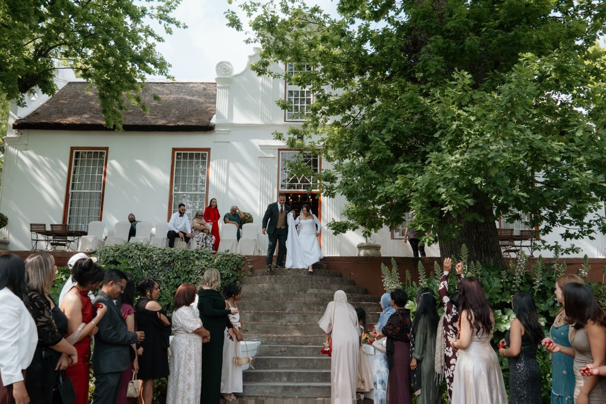 Wedding guests gather on stone steps outside white Cape Dutch venue as bride and groom exit ceremony.