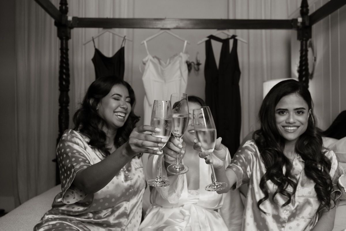 Black and white photo of bridesmaids in robes toasting champagne on a bed with wedding dresses hanging behind them.
