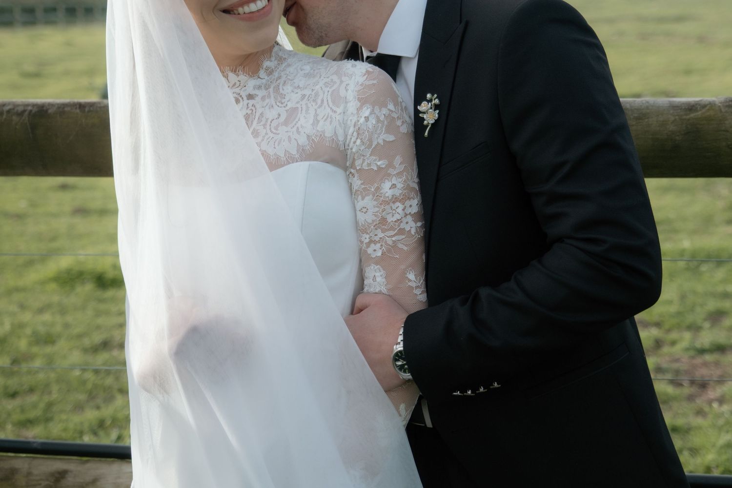 A groom in a black tuxedo kisses his bride in a lace wedding dress and veil outdoors on a farm.