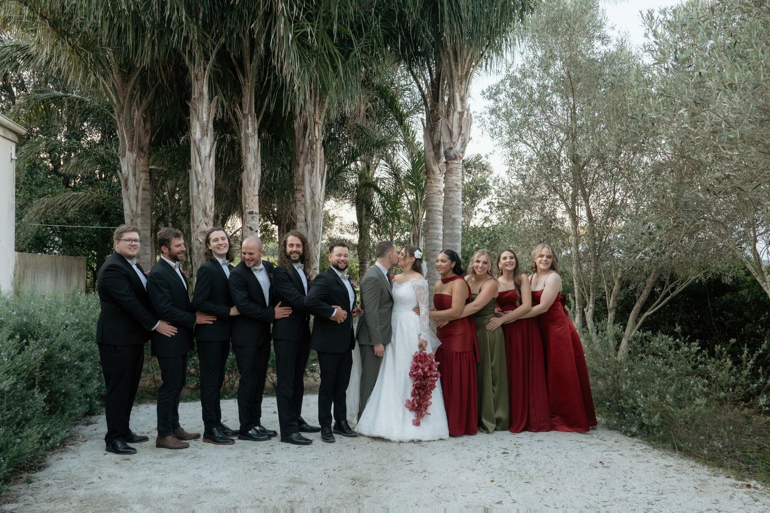 Wedding party poses outdoors among palm trees, groomsmen in black suits and bridesmaids in red gowns flanking the bride and groom.