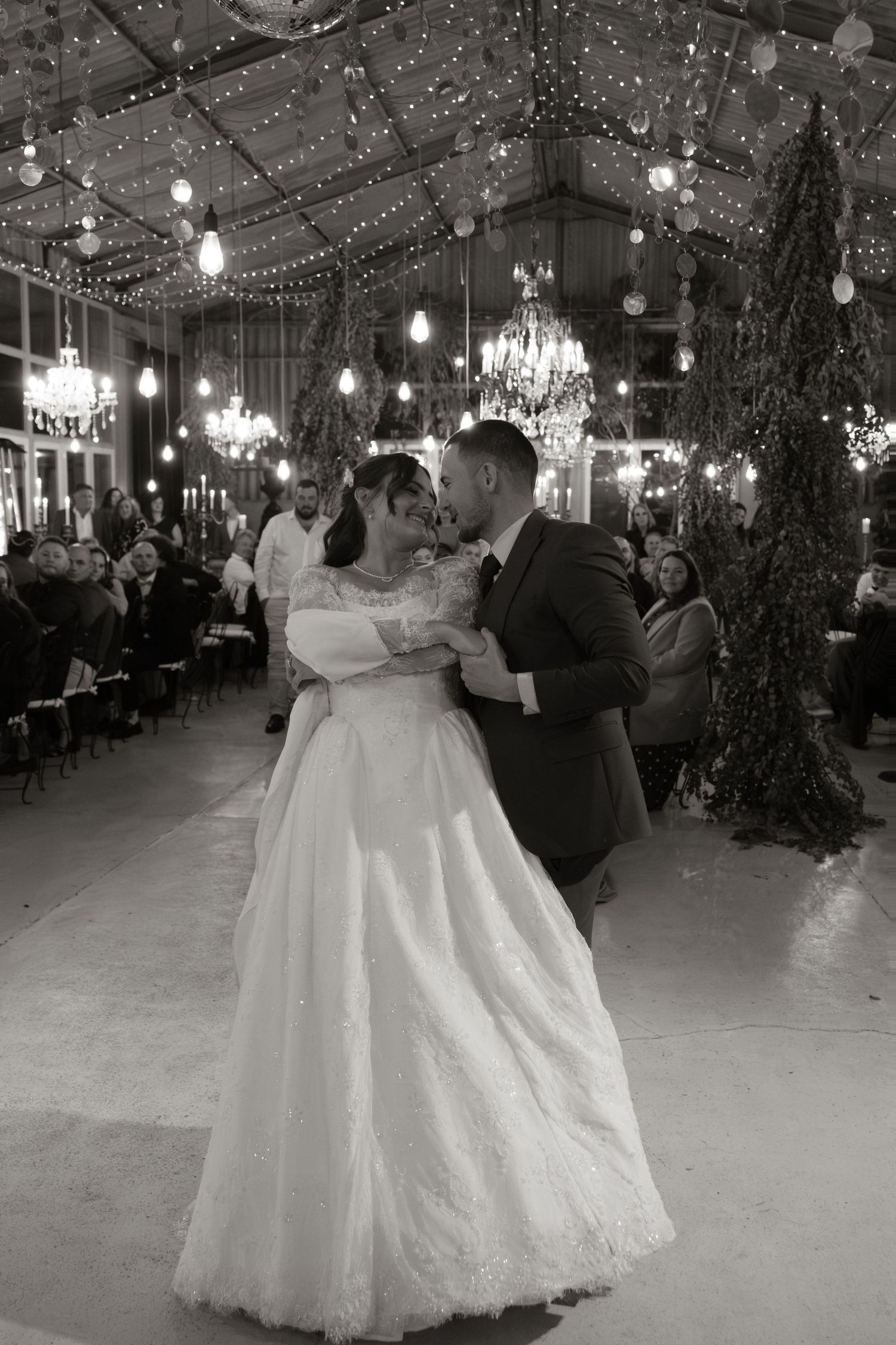 Bride and groom share first dance in black and white photo under chandeliers and string lights.