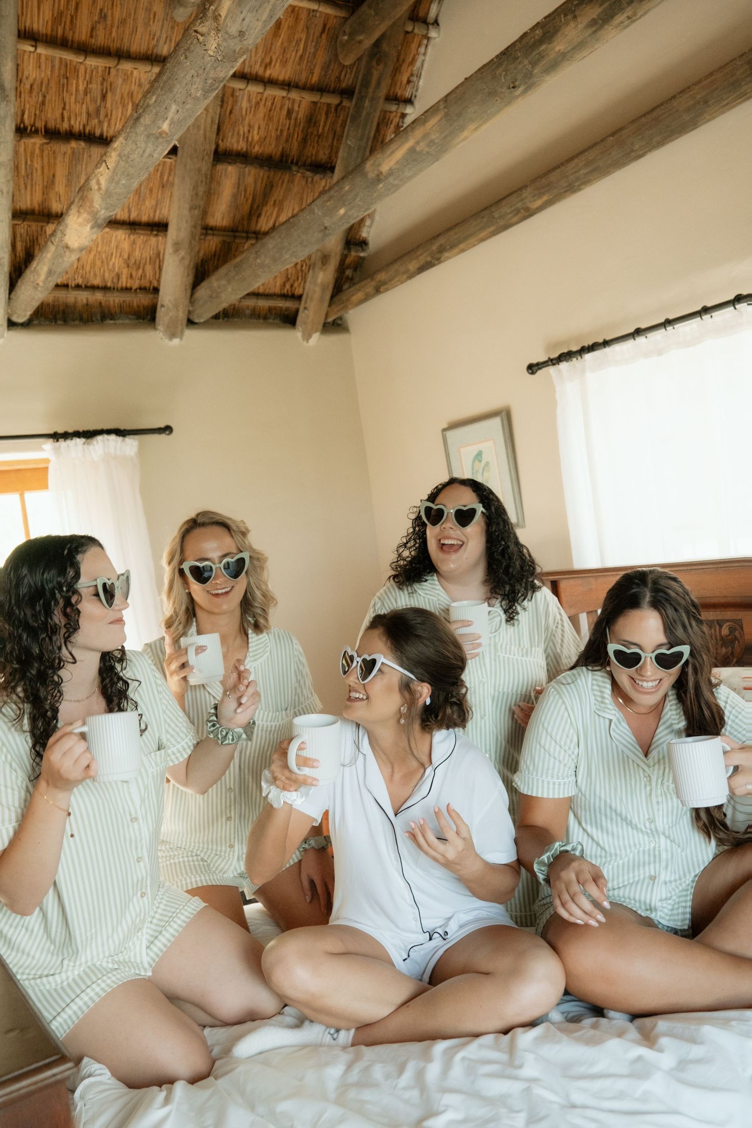 Bridesmaids in pajamas and heart sunglasses enjoy coffee mugs on a bed before the wedding.