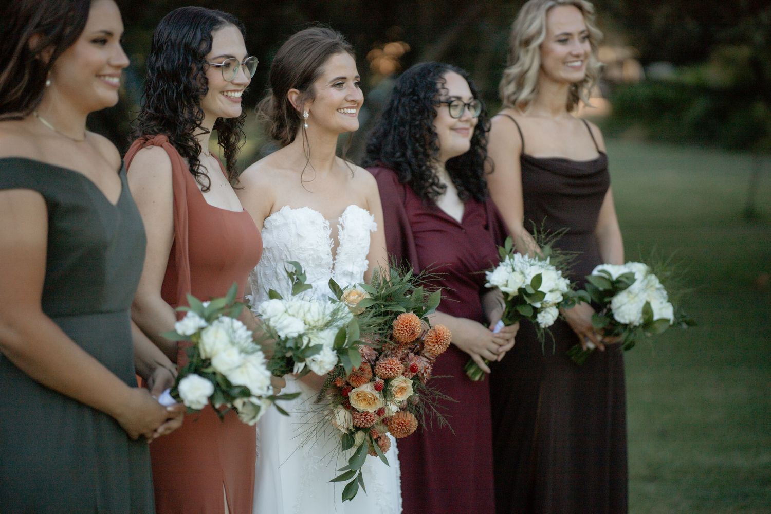 Bride poses with five bridesmaids in mismatched earth-tone dresses holding white and orange bouquets.