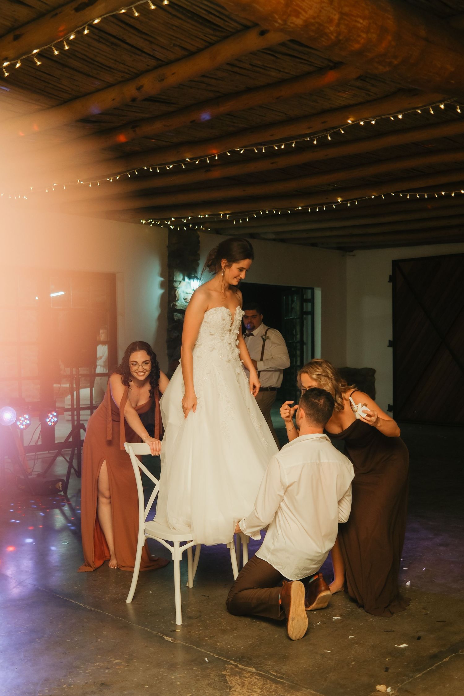 Bride standing on chair in white gown assisted by bridesmaids and groom at rustic wedding venue with fairy lights.