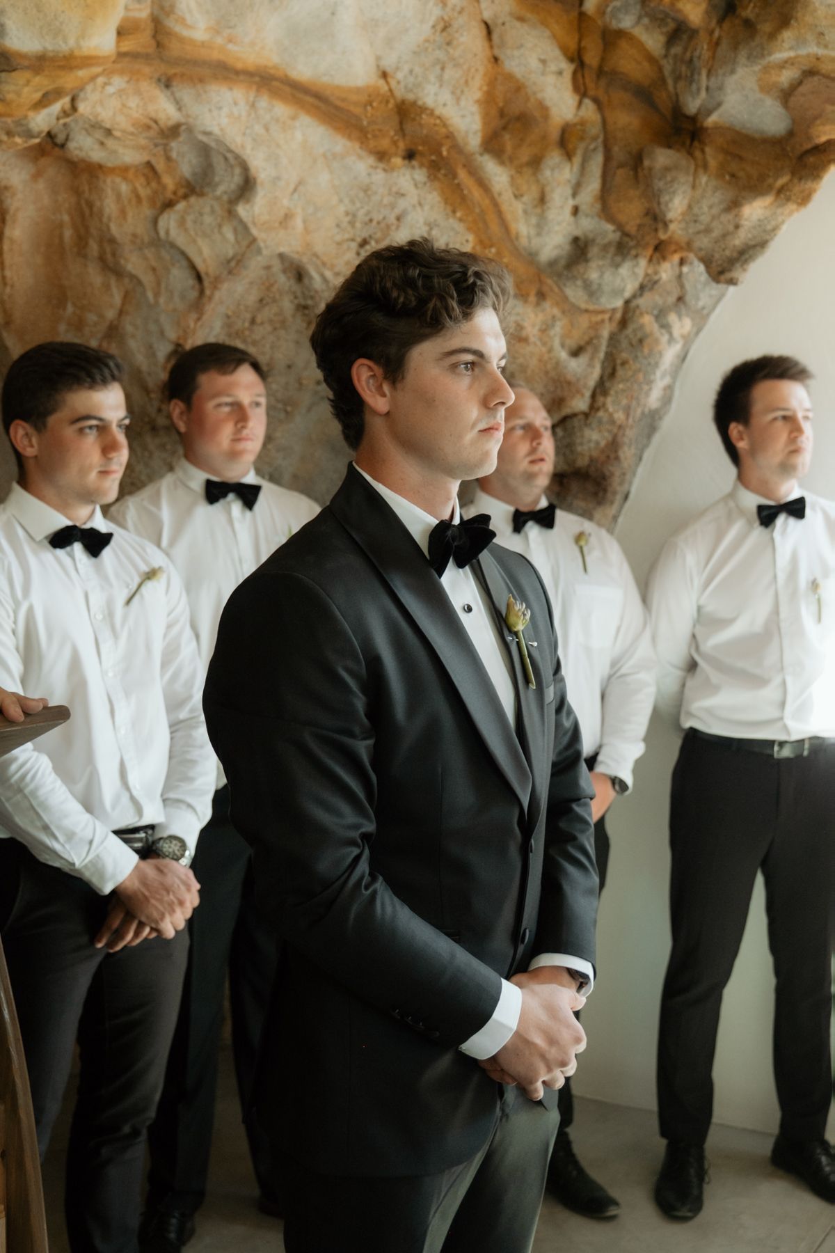 Groom in black tuxedo with boutonniere stands before groomsmen in white shirts and bow ties at elegant wedding ceremony.