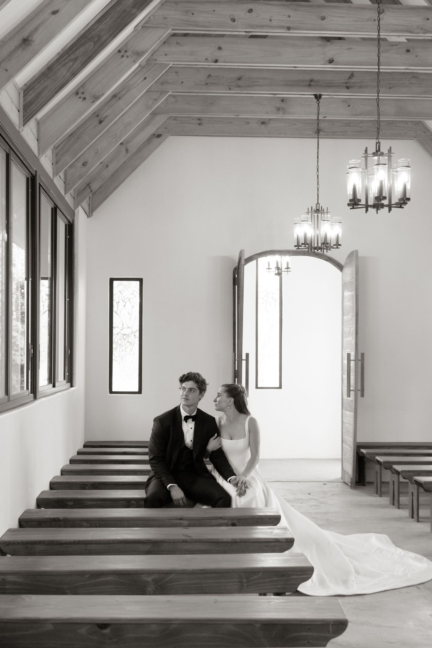 Black and white photo of bride and groom sitting on chapel pew with vaulted wood ceiling and chandeliers.