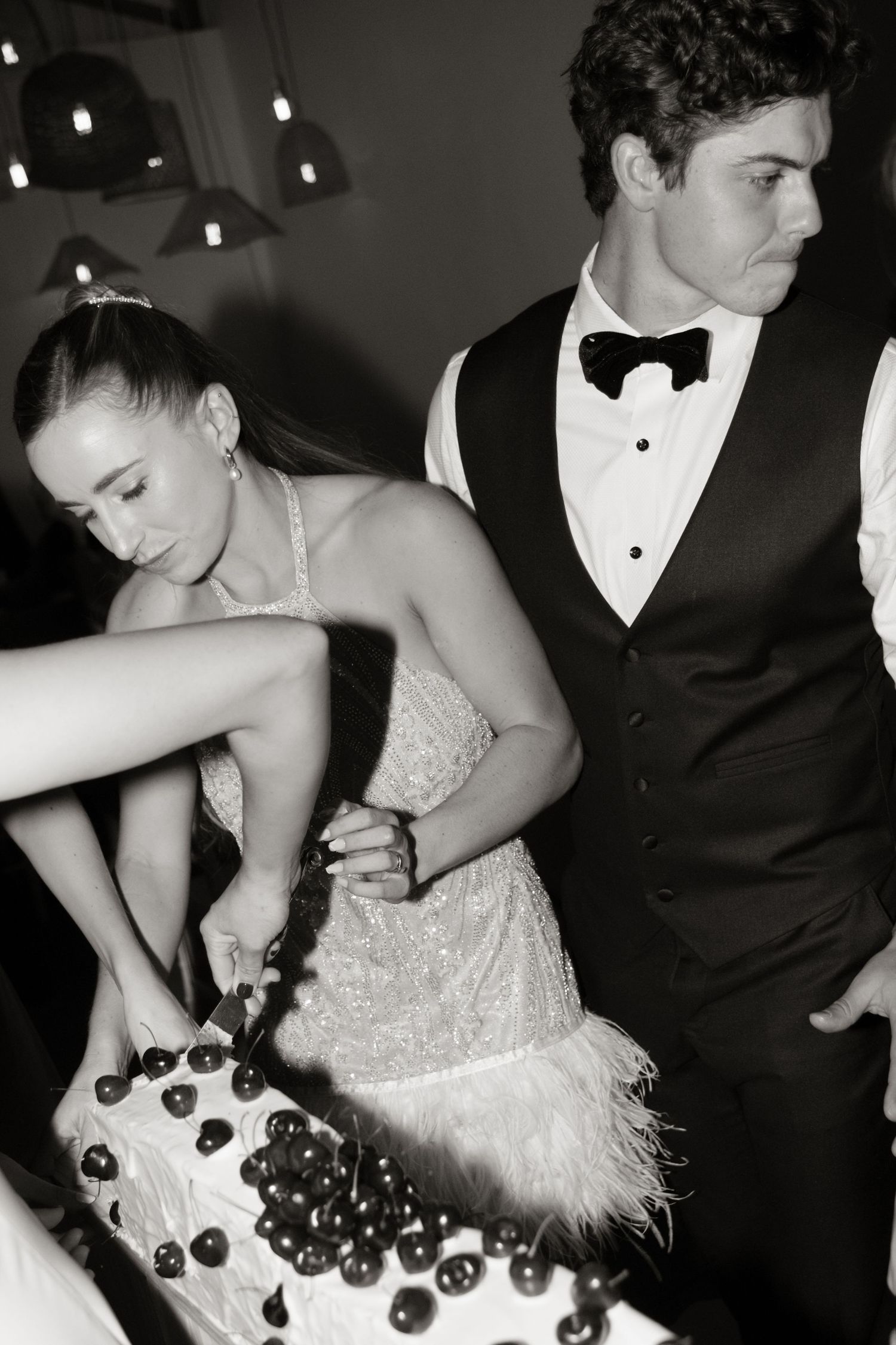 Black and white photo of bride and groom cutting cherry-topped wedding cake at reception.