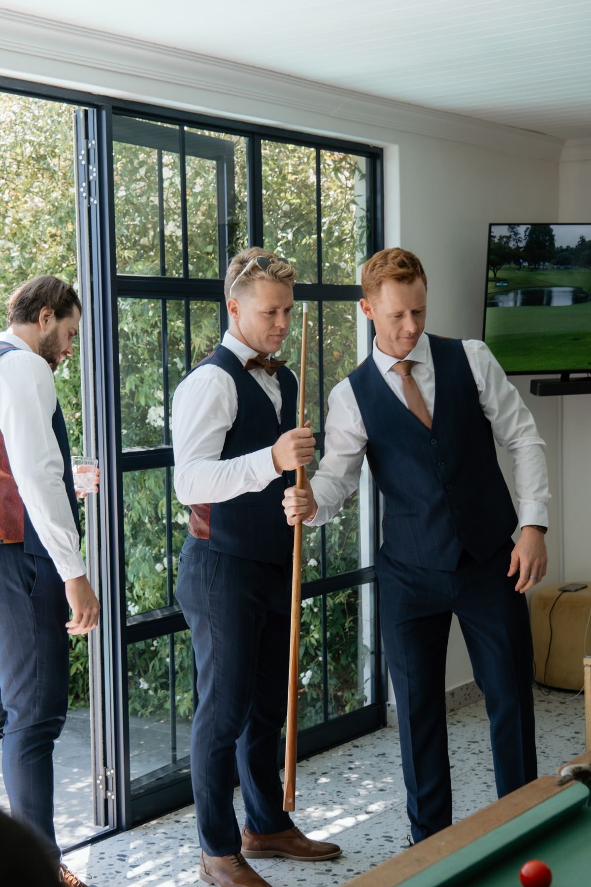 Groomsmen in navy vests play pool indoors before a wedding ceremony near large windows.