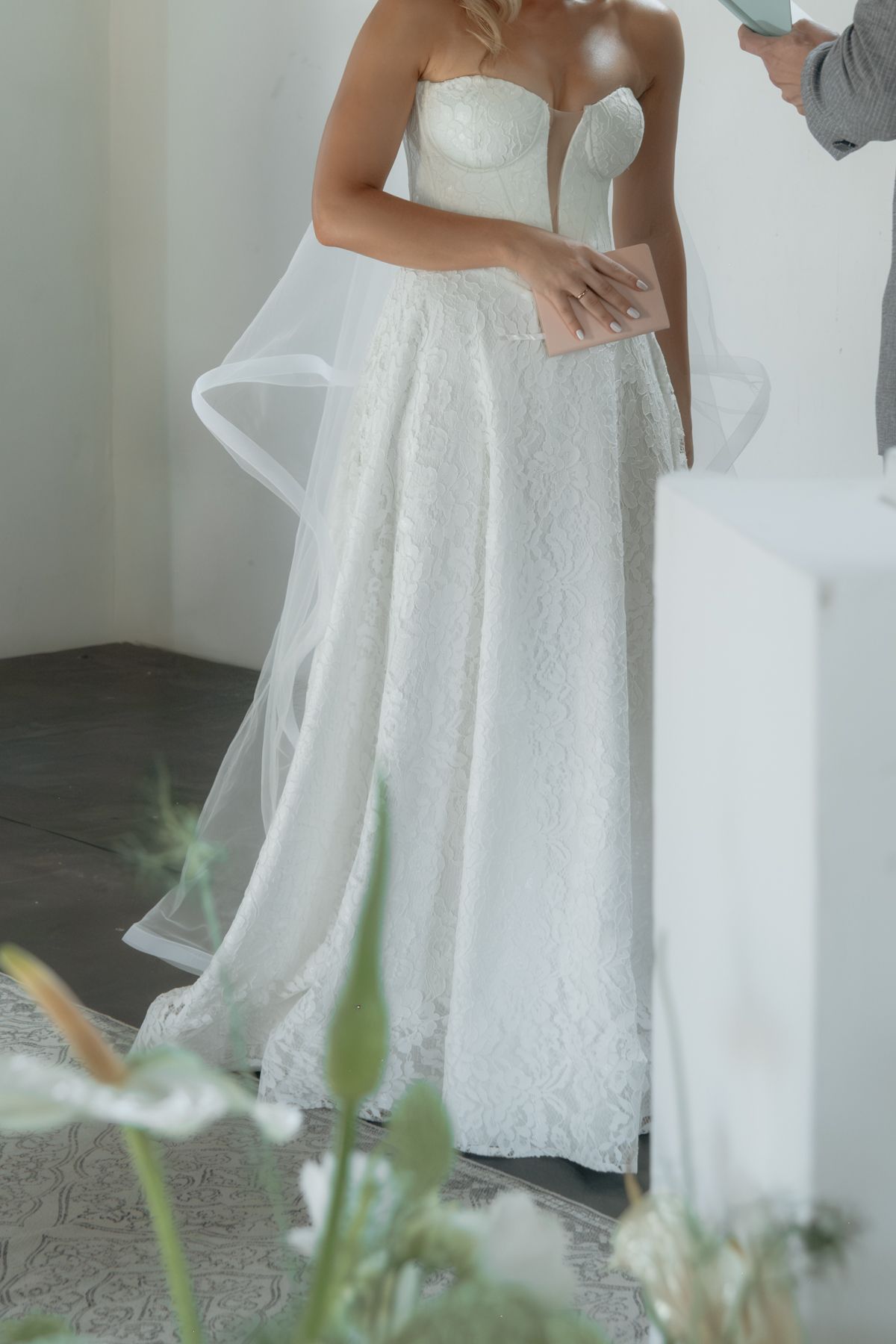 Bride in a strapless lace wedding gown with tulle overlay being assisted in a bright white room.