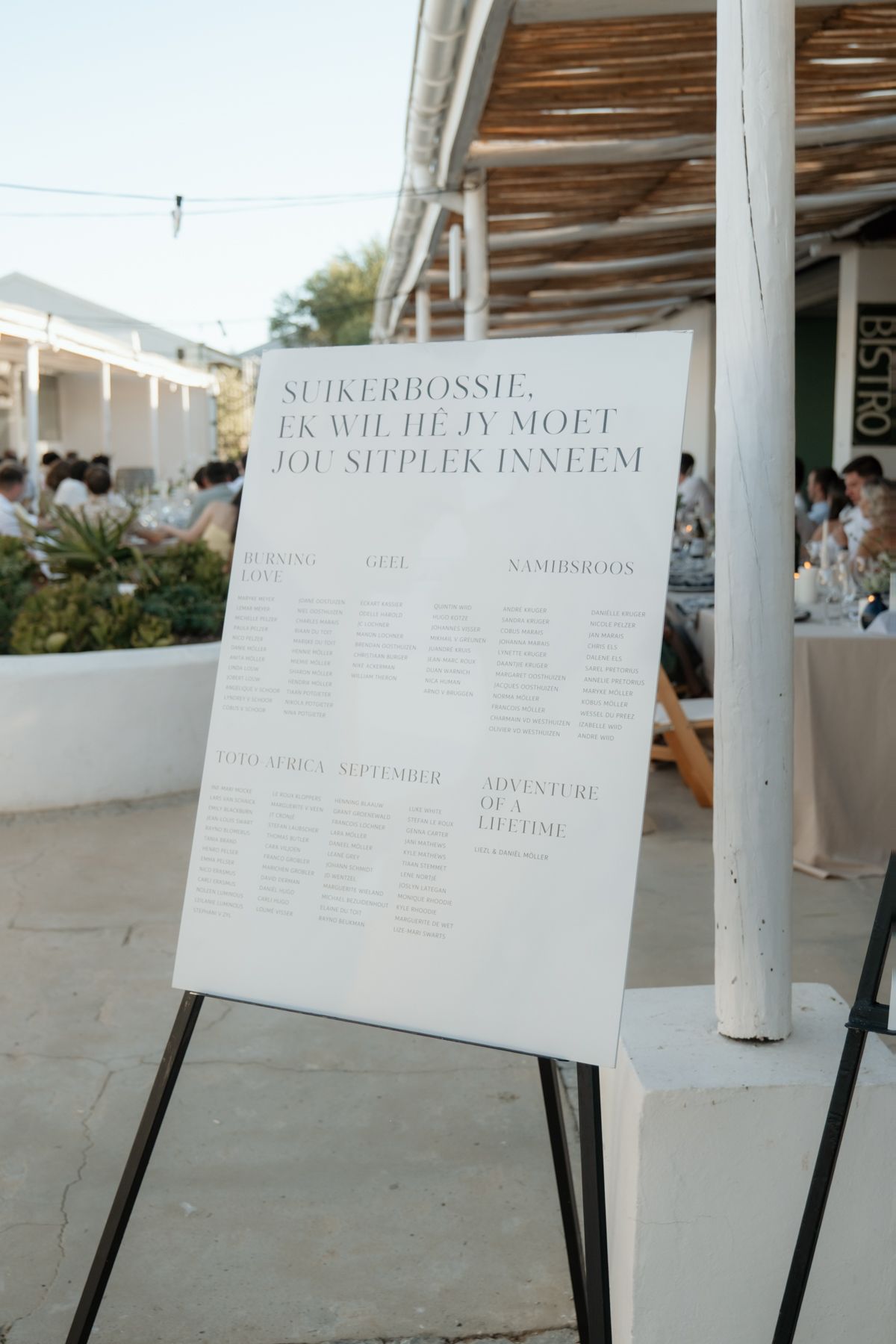Elegant white wedding seating chart sign on easel with Afrikaans text at outdoor reception venue.