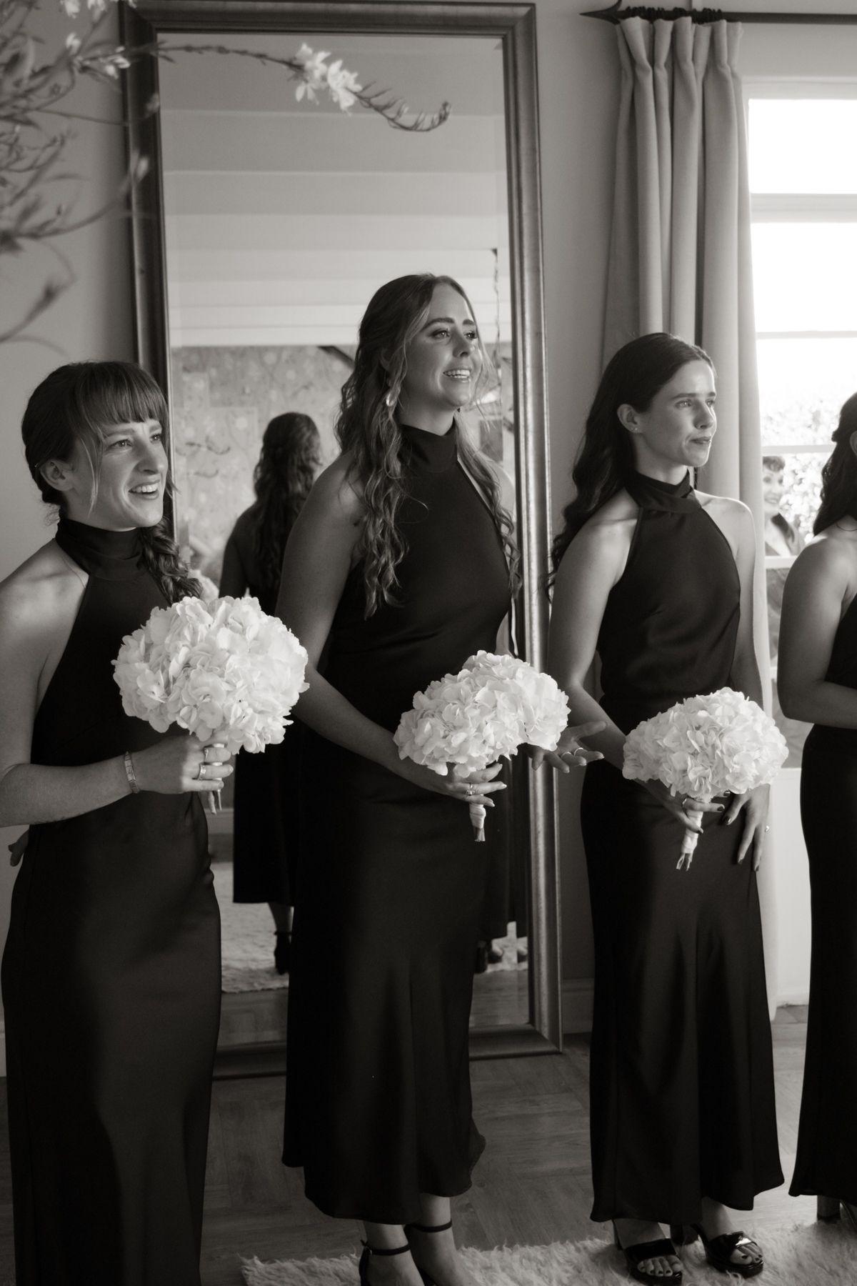 Three bridesmaids in black halter dresses holding white hydrangea bouquets in a black and white photo.