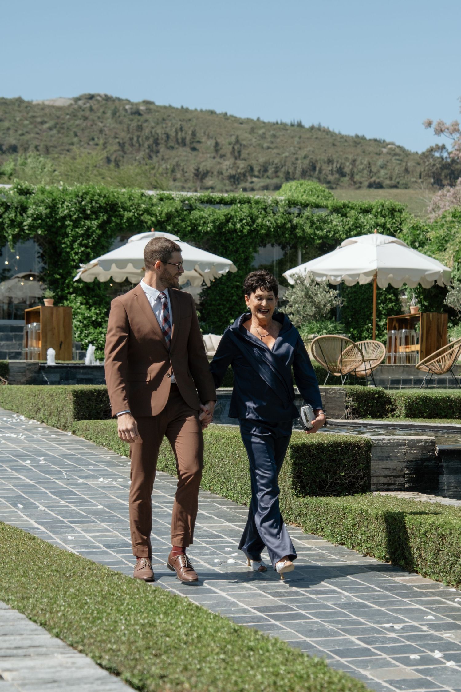 Groom in brown suit walks with a woman in navy outfit through manicured garden with mountain backdrop on wedding day.