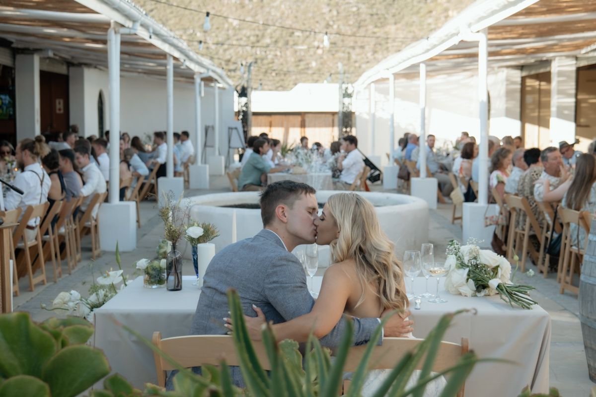 Newlyweds sharing a kiss at their reception table with guests seated in elegant open-air venue.