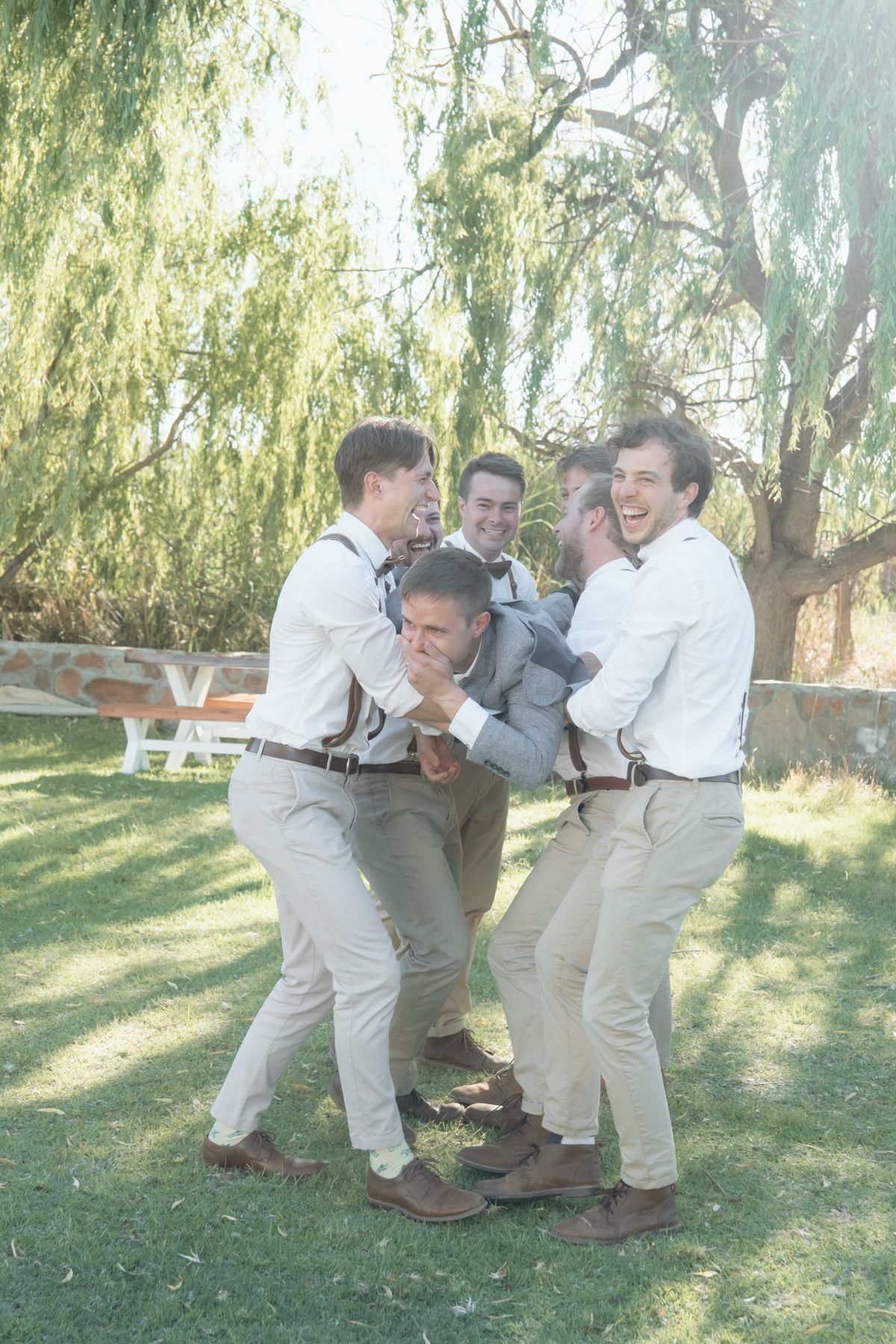 Groomsmen in white shirts and khakis playfully lifting groom in grey suit under a willow tree.