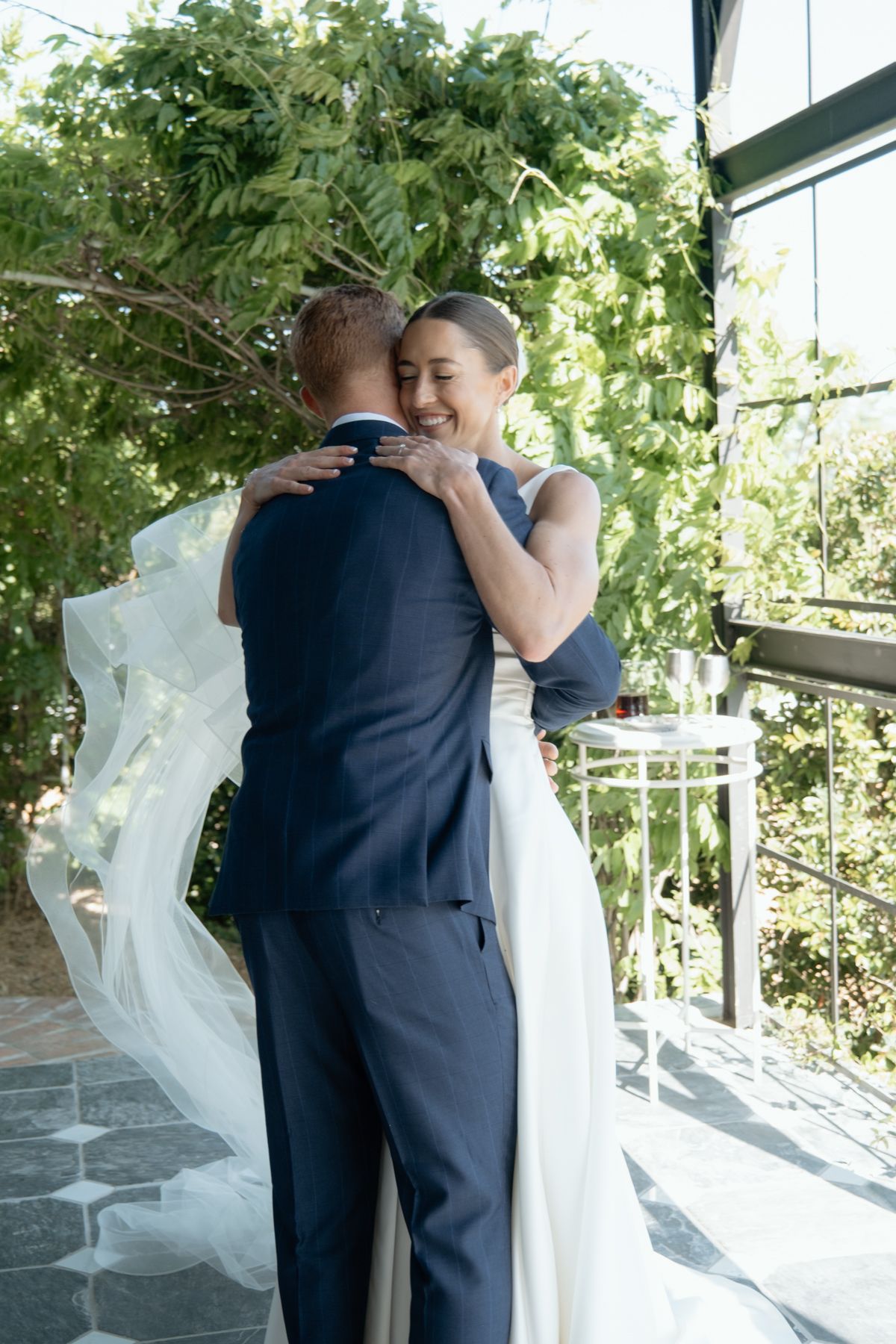 Smiling bride in white gown with veil embraces groom in navy suit during outdoor wedding ceremony.